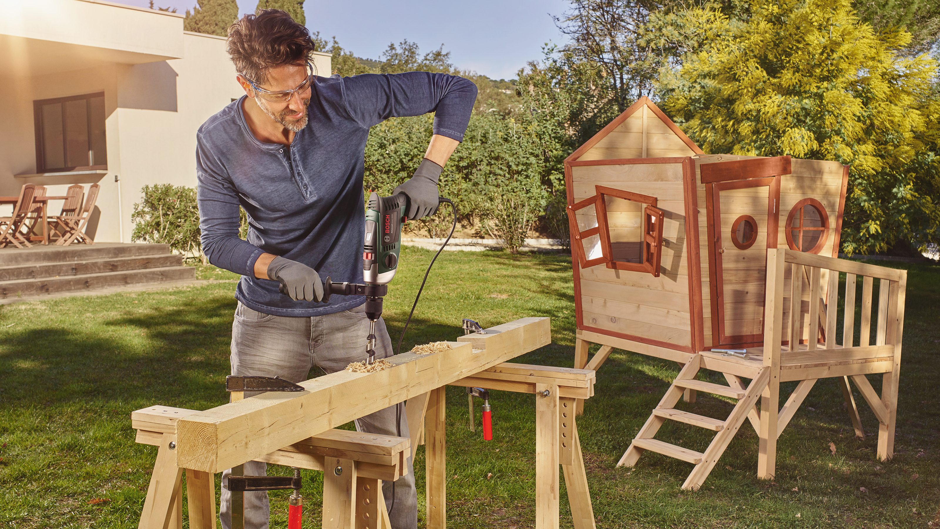 Mężczyzna wierci w drewnie przed domkiem na drzewie w ogrodzie. Słońce oświetla scenę.