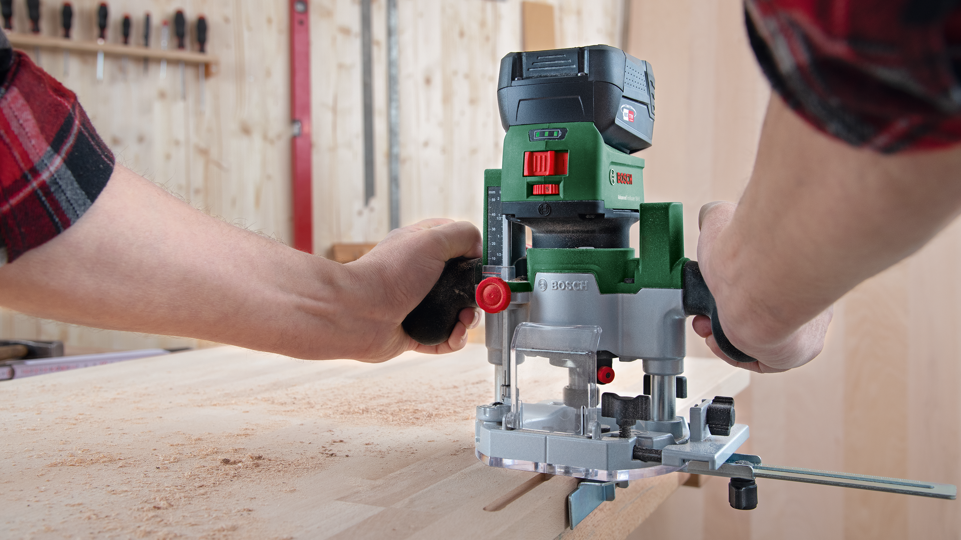 Mains guidant une défonceuse Bosch verte sur du bois. Outils accrochés en arrière-plan.