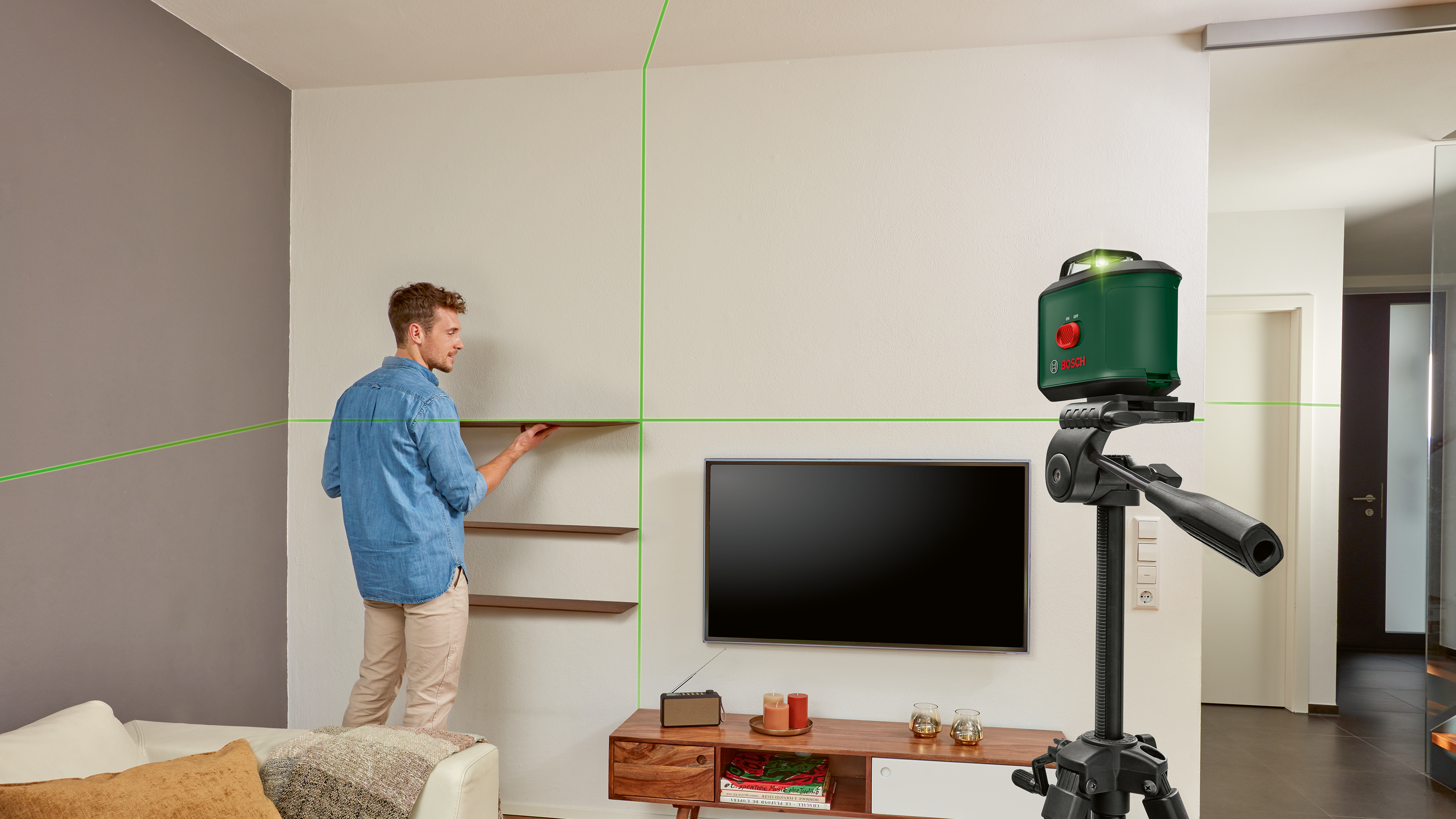 Mężczyzna montuje półkę, niwelator laserowy na statywie. Salon z TV.