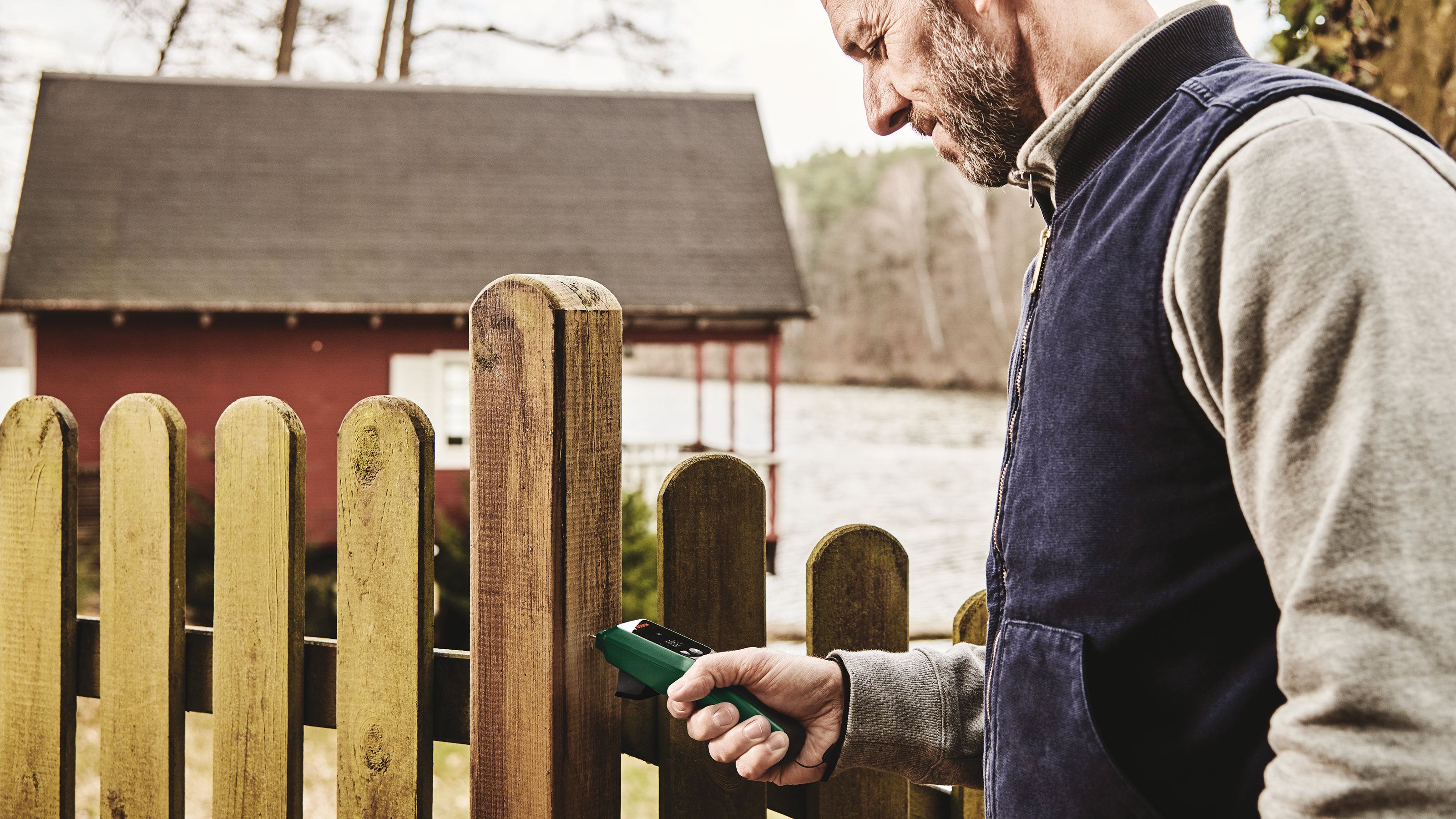 Homme barbu utilise appareil vert sur clôture en bois près maison rouge.