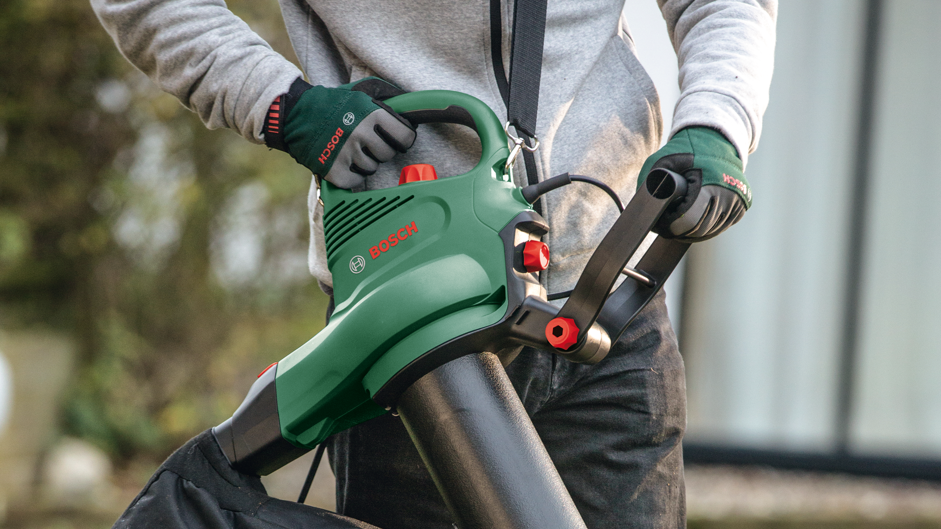 Mężczyzna w szarej bluzie i zielonych rękawicach trzyma zielony dmuchawę.