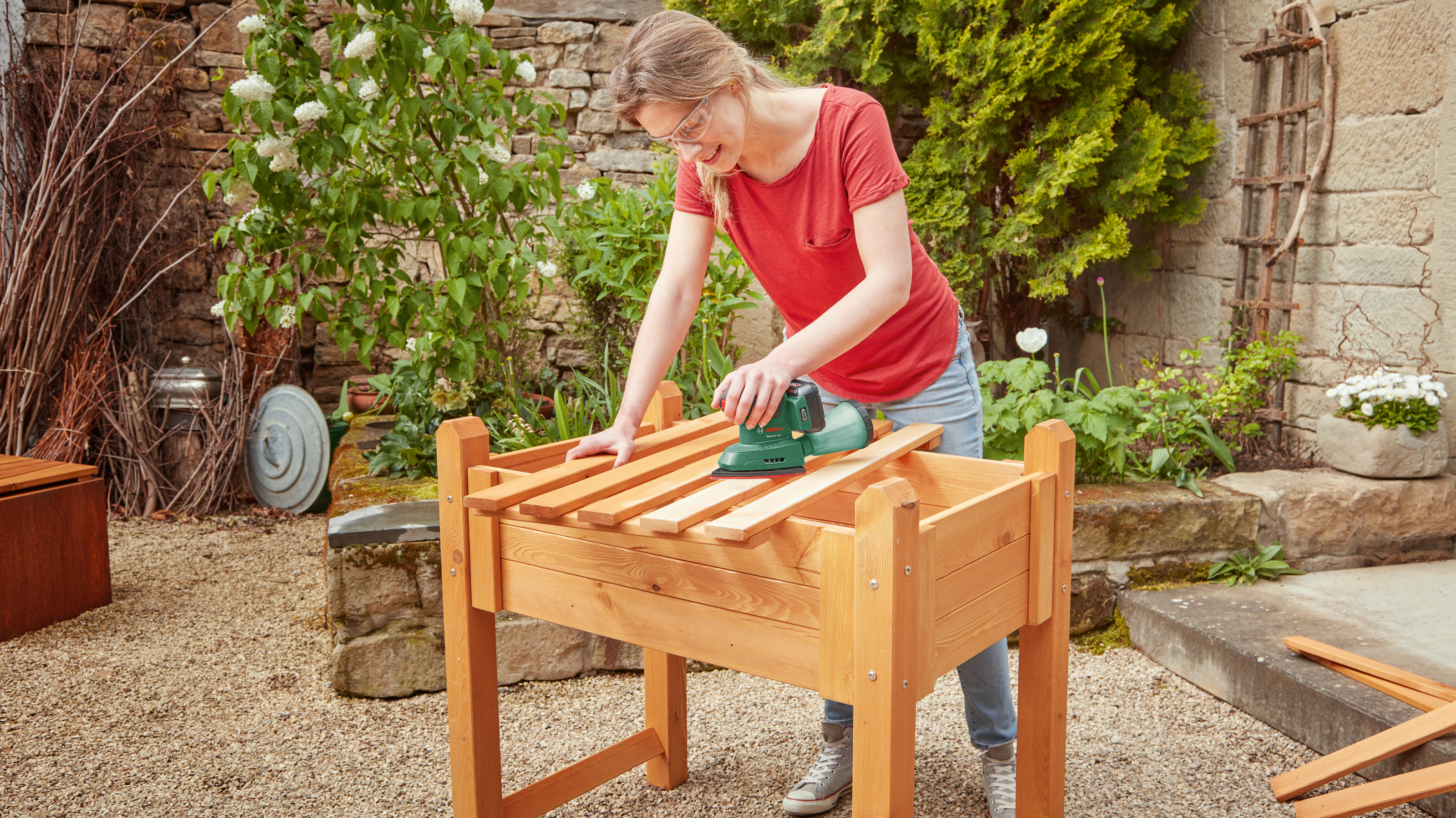Femme ponçant un cadre de jardinière surélevée en bois avec une ponceuse verte.