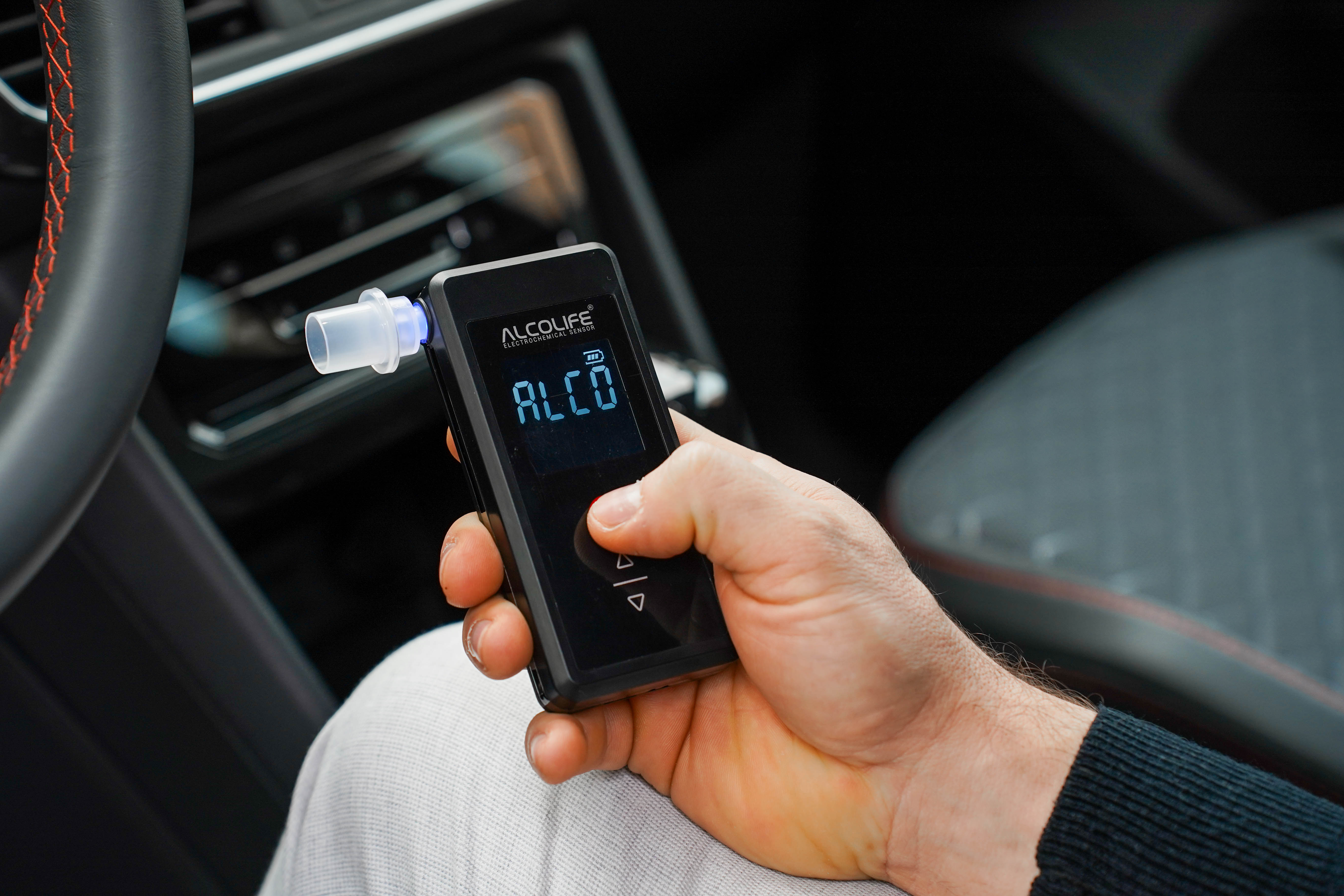 Ręka trzyma czarny alkomat z niebieskim wyświetlaczem w samochodzie.