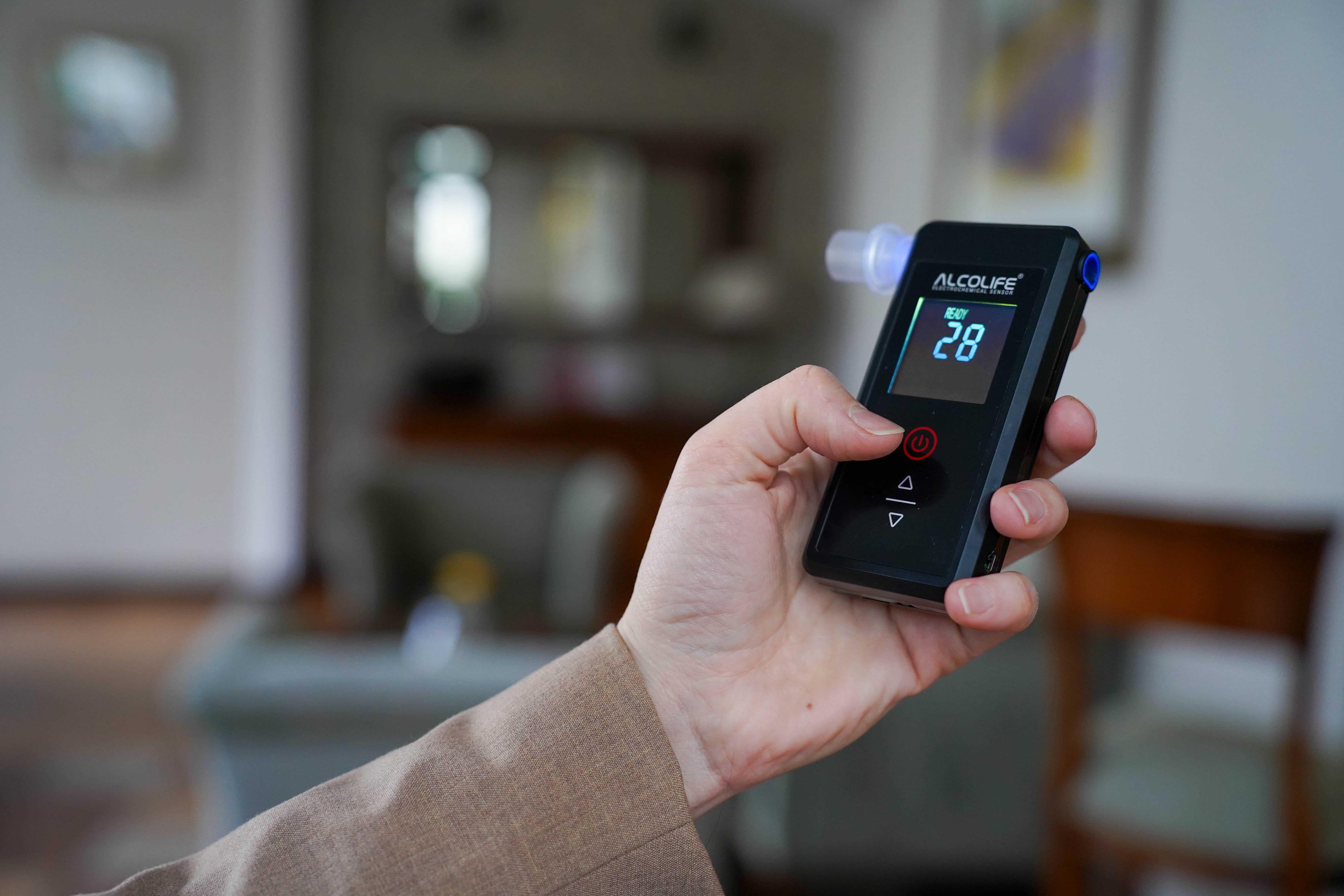 Dłoń trzyma czarny alkomat z niebieskim wyświetlaczem pokazującym '28'.