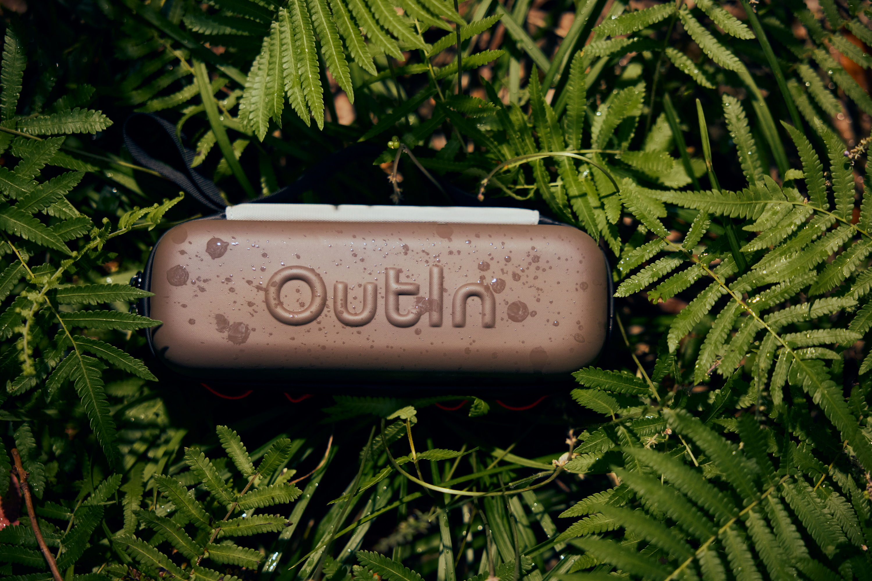 Een bruine reistas met 'Outlin' logo tussen groene varens.