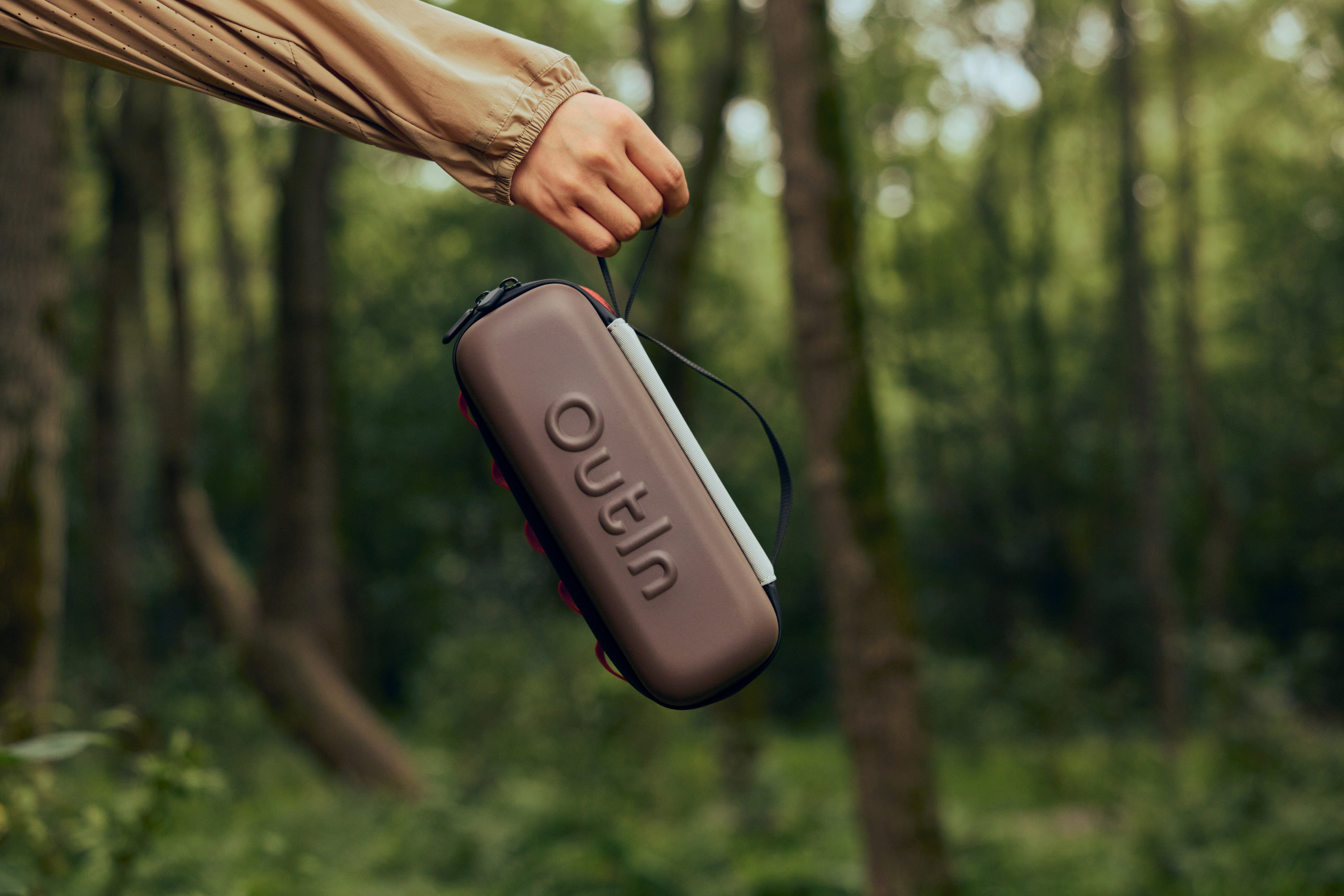 Een hand houdt een bruine tas met "Outin" logo vast voor bosachtergrond.