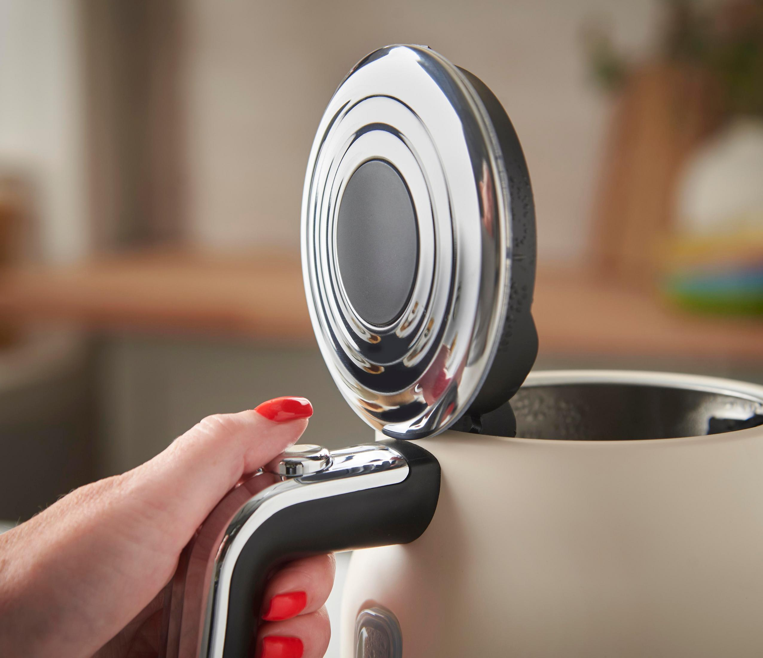 Eine Hand mit roten Nägeln öffnet den Deckel eines cremefarbenen Wasserkochers.