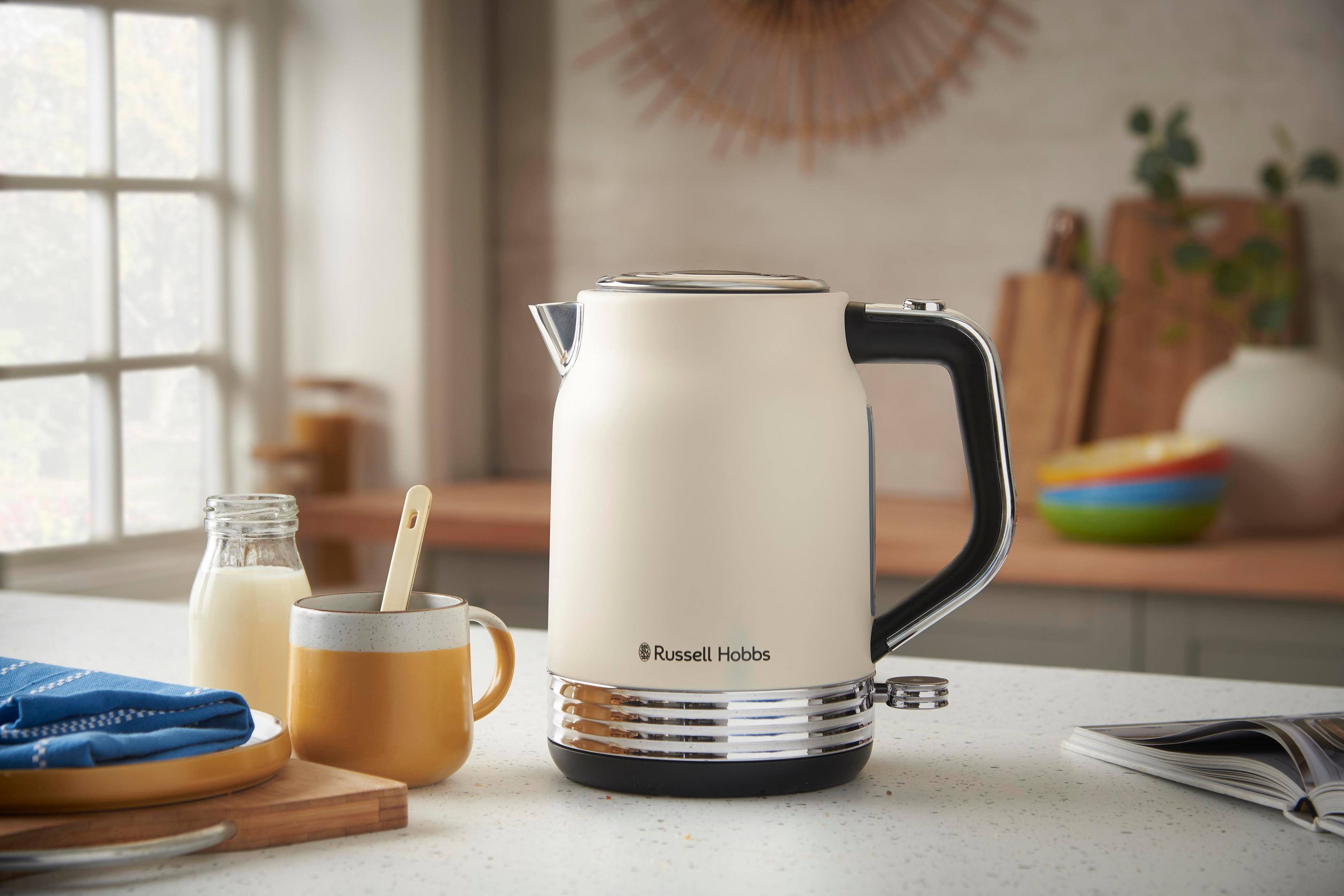 Eine cremefarbene Russell Hobbs-Kanne mit schwarzem Griff steht auf einer Arbeitsplatte neben einer Tasse.
