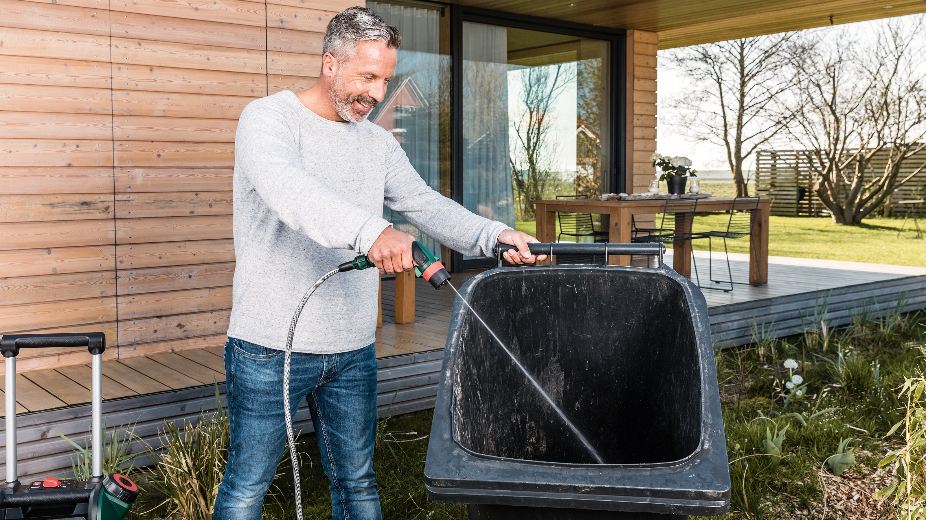 Homme nettoie poubelle avec tuyau d'arrosage devant maison bois.