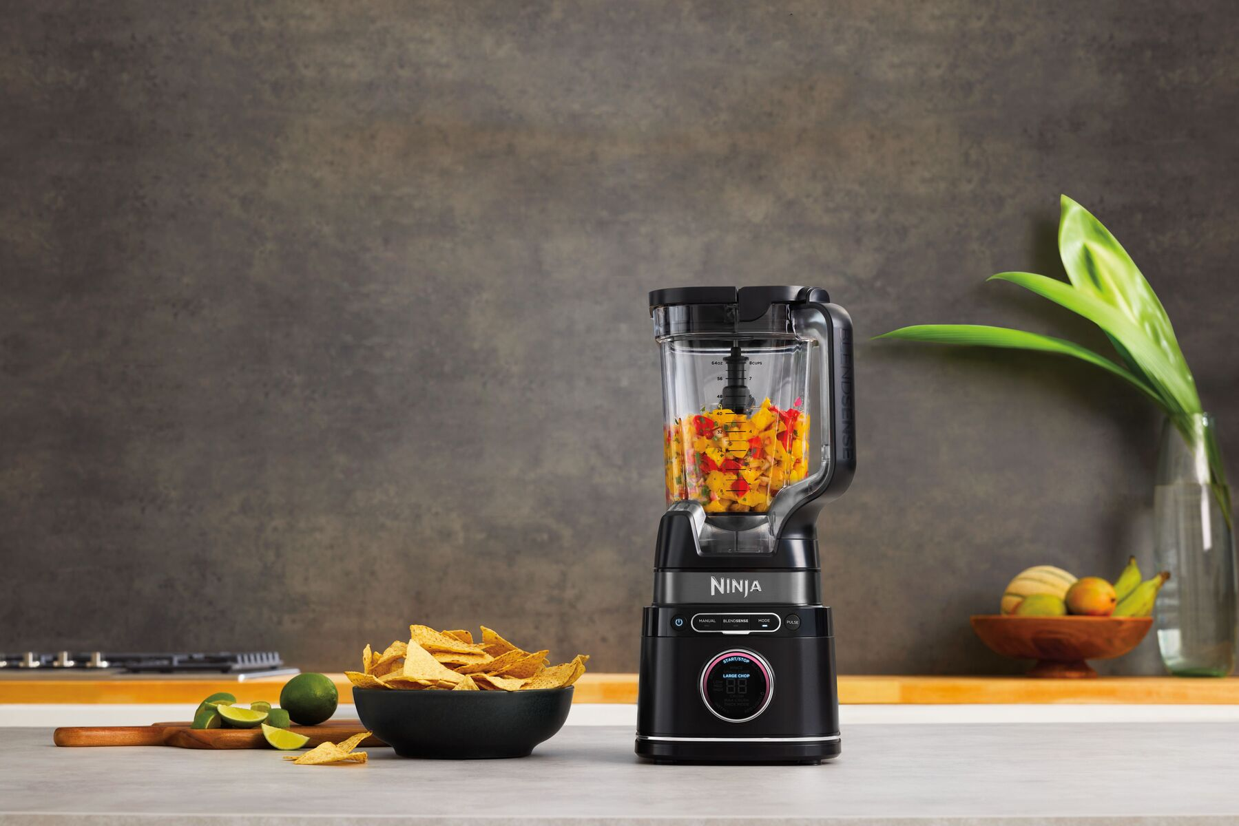 A black Ninja blender with chopped vegetables, next to a bowl of chips and limes.