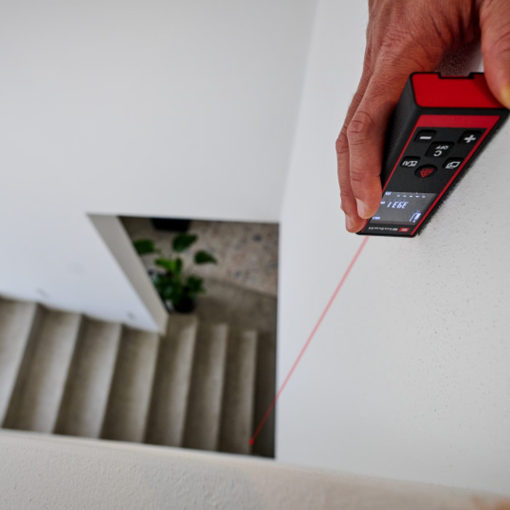 Eine Hand hält ein rotes Messgerät, das einen roten Laserstrahl auf eine weiße Wand projiziert.