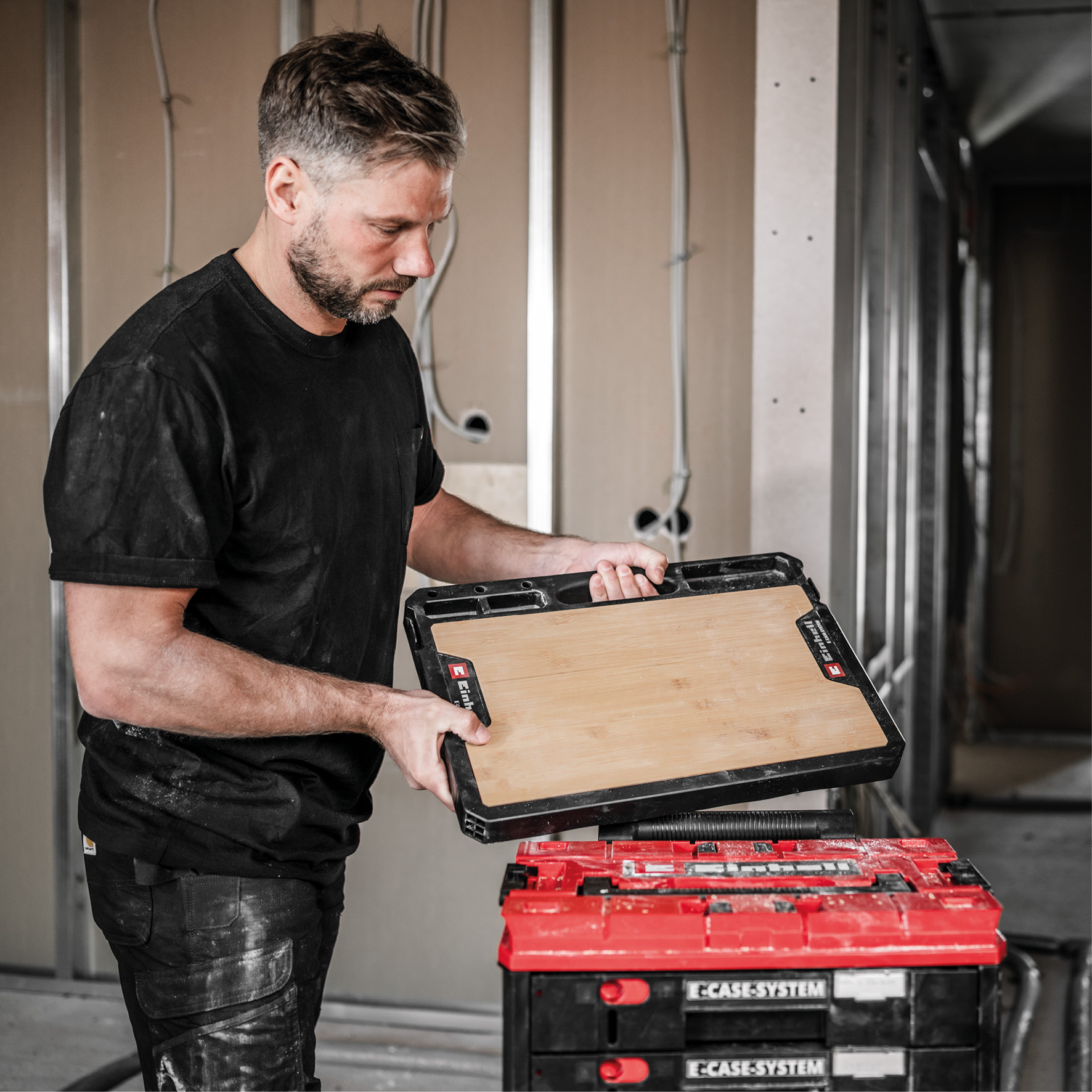 Ein Mann in Arbeitskleidung hält eine Holzplatte über rote Werkzeugkisten.
