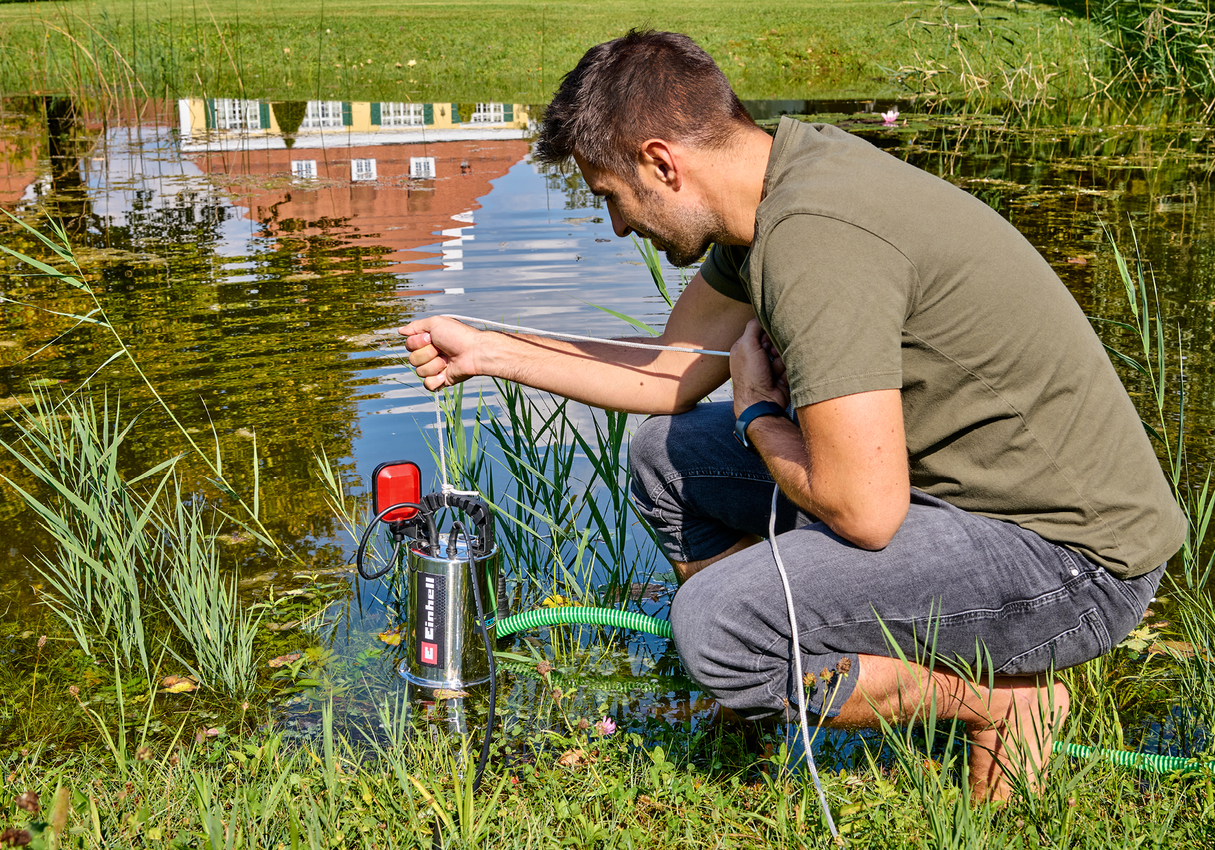 Mann mit Tauchpumpe im Teich, reflektiertes Gebäude im Hintergrund.