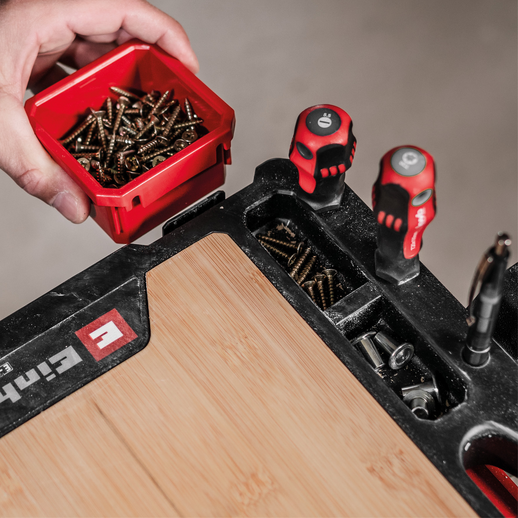 Eine Hand hält einen roten Schraubenbehälter über eine Werkzeugbox mit Schraubendrehern.