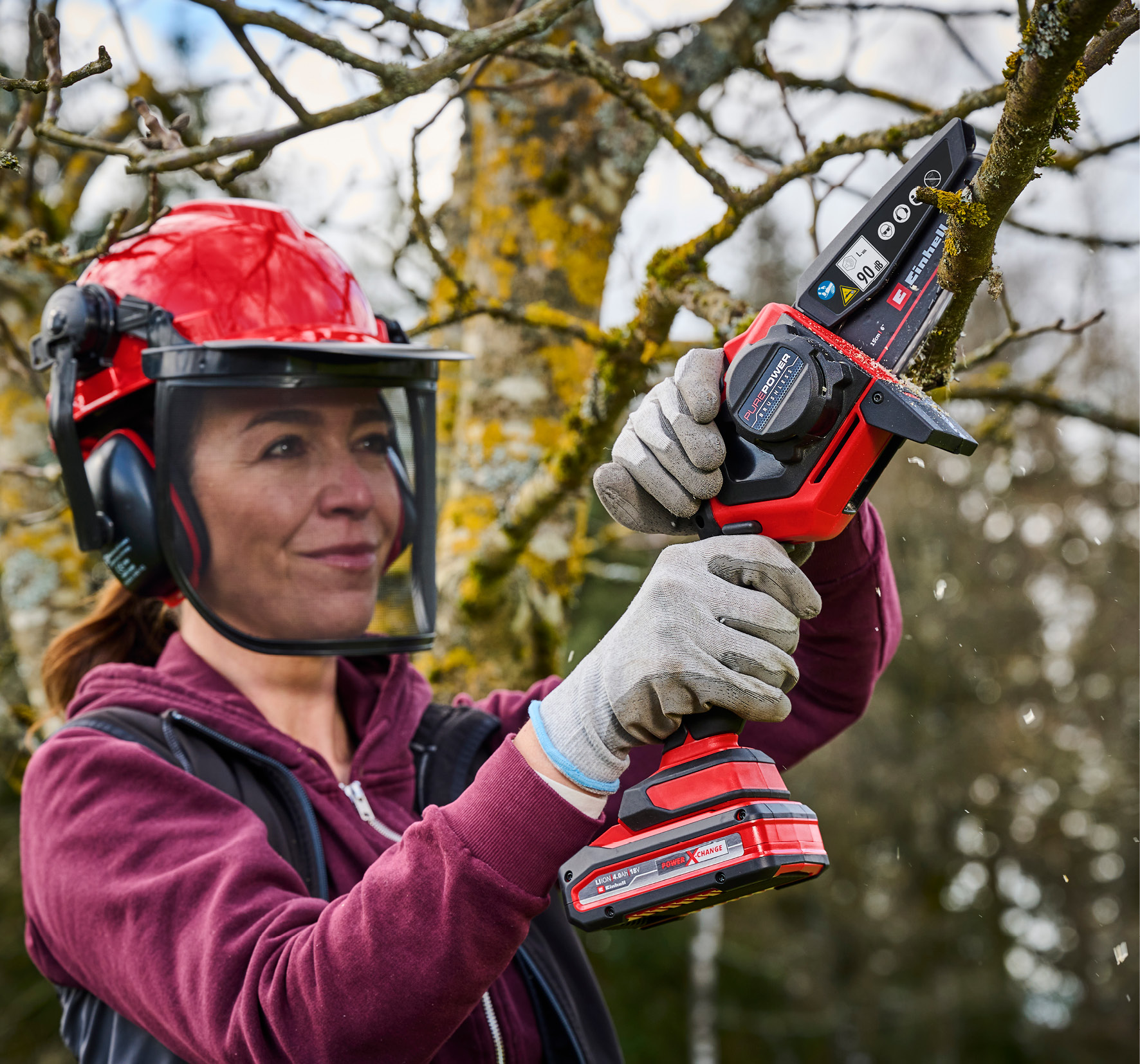 Eine Frau mit Helm und Schutzbrille benutzt eine Kettensäge an einem Baum.