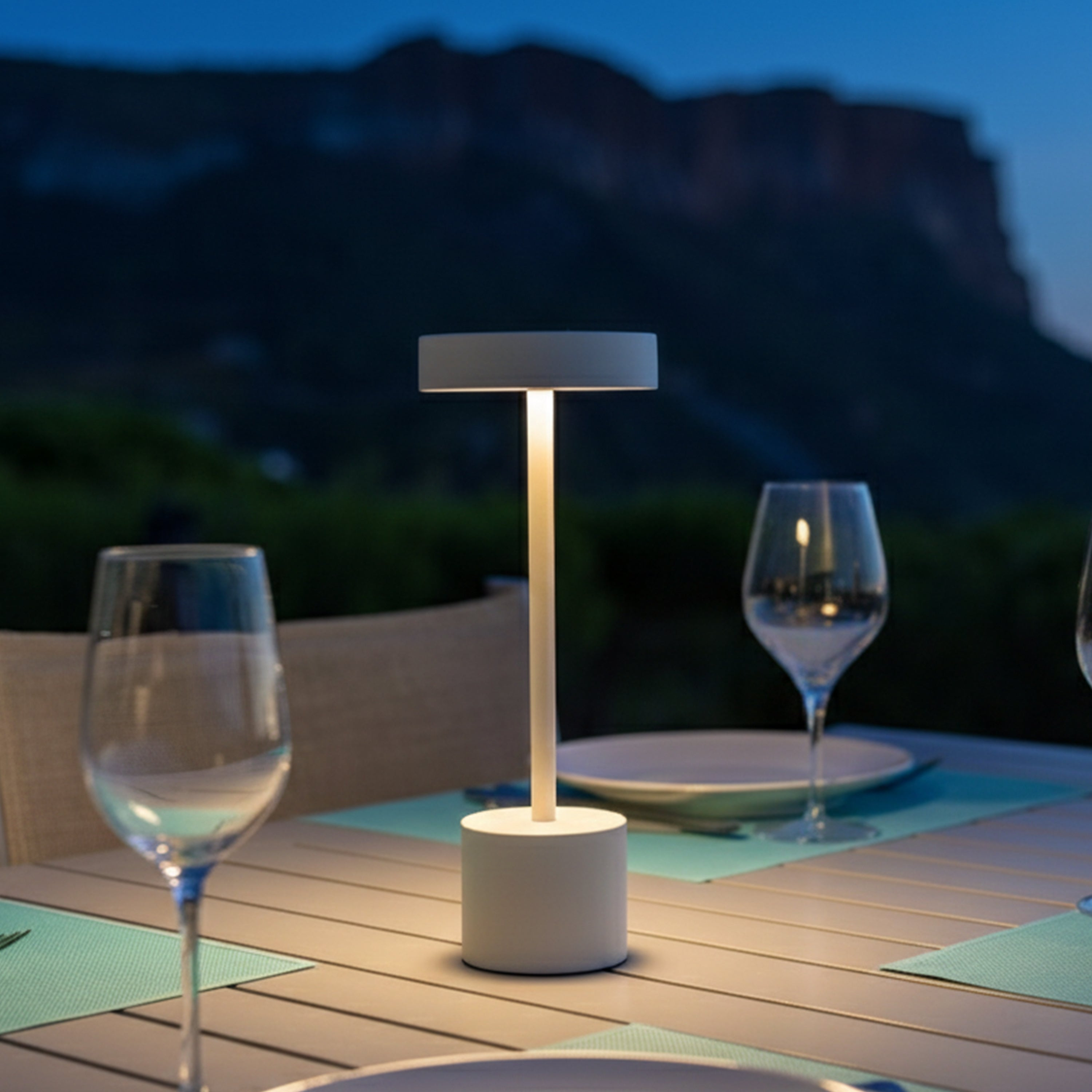 Eine weiße Lampe beleuchtet einen Tisch mit Gläsern und Tellern im Freien bei Dämmerung.