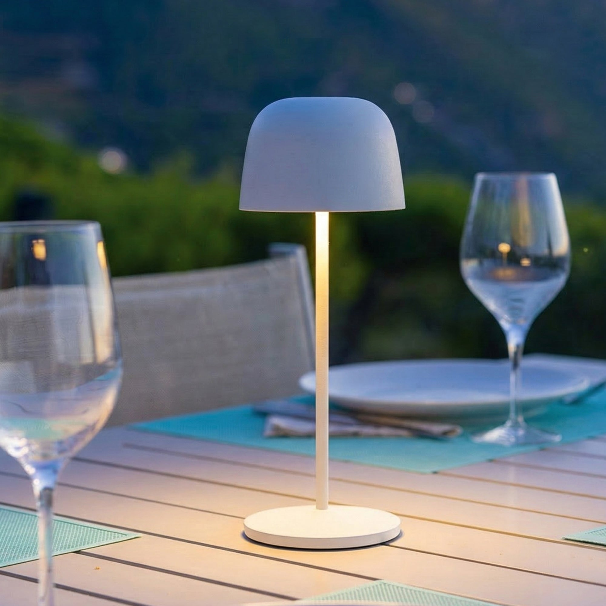 Eine weiße Tischlampe steht auf einem Tisch mit Gläsern und Tellern im Freien.