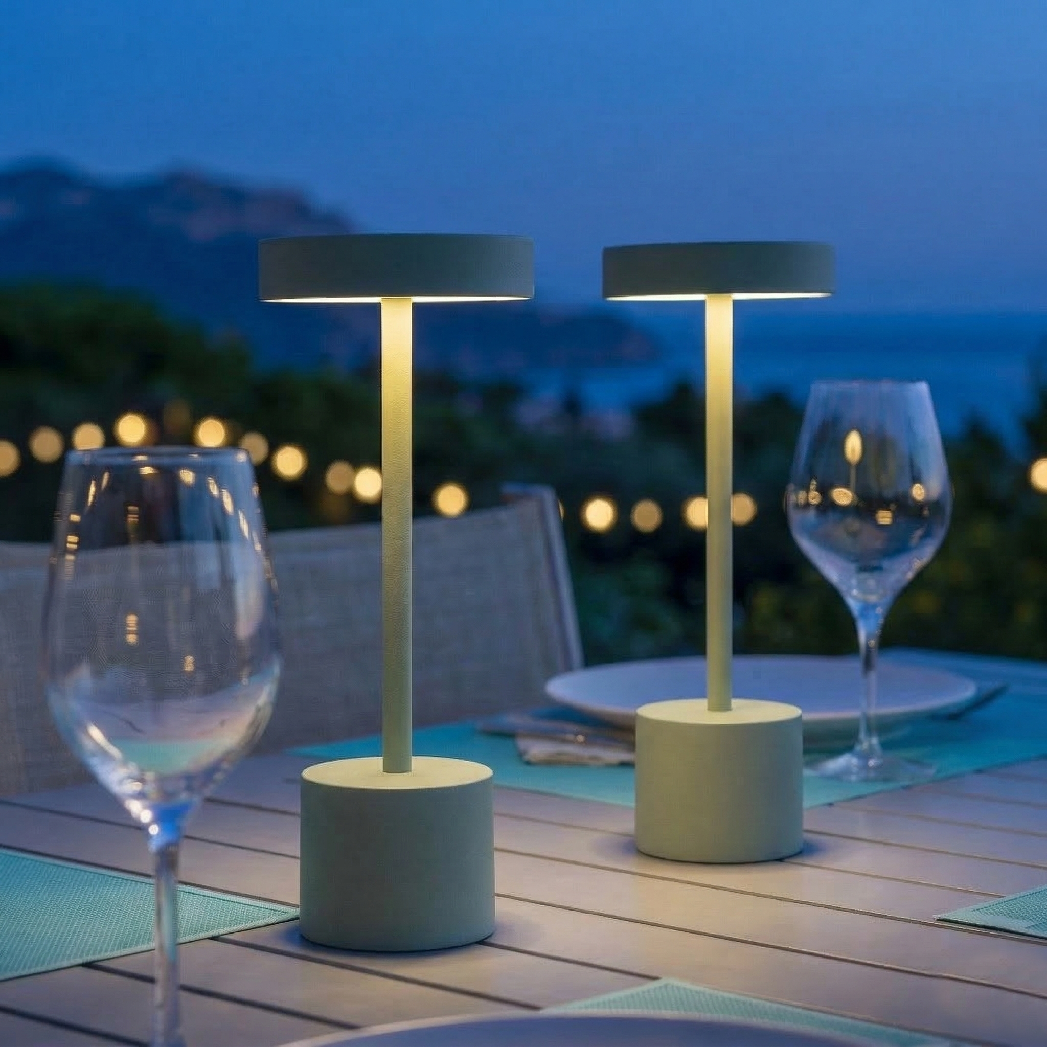 Zwei Tischlampen auf einem Tisch im Freien bei Dämmerung mit Lichtern im Hintergrund.