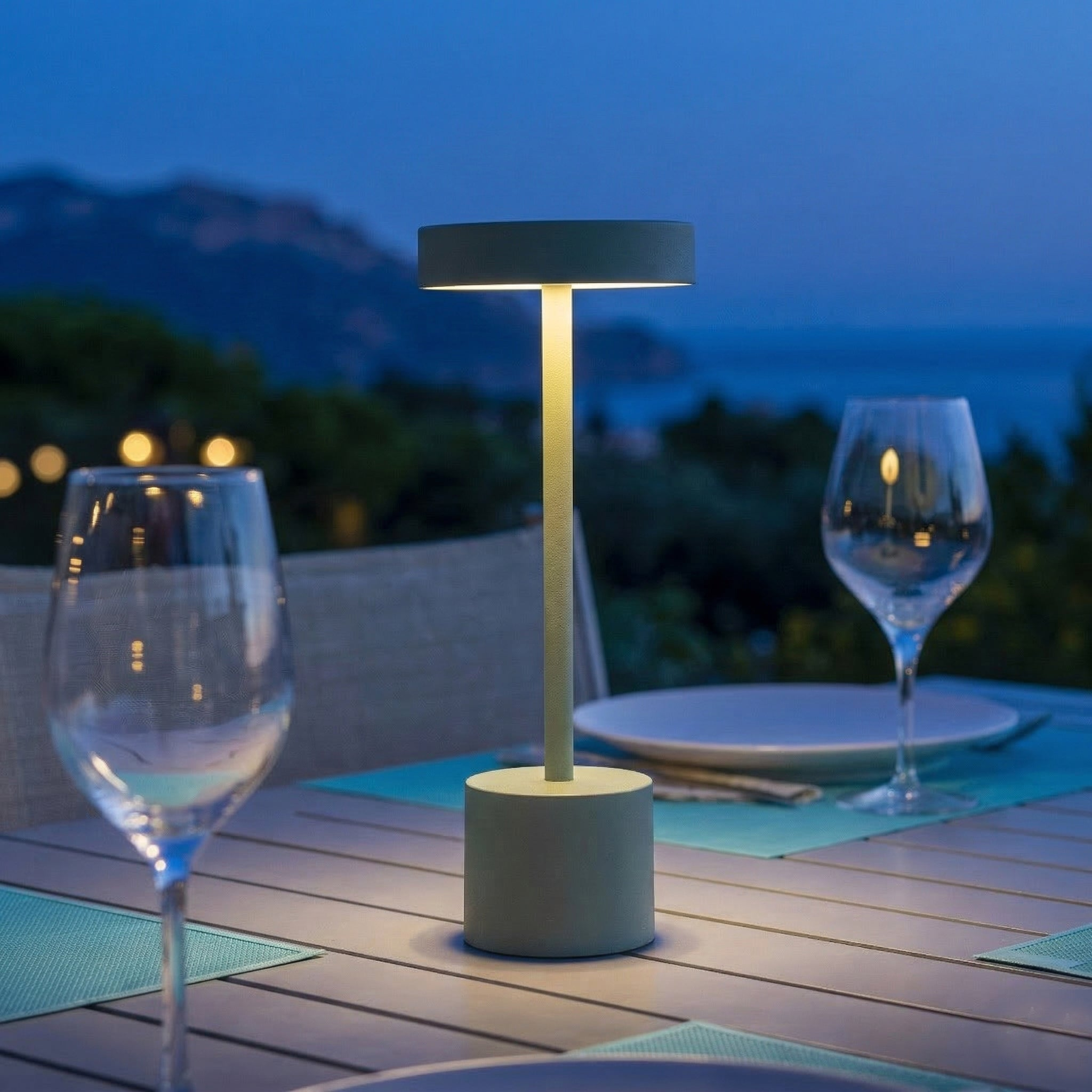 Eine Tischlampe beleuchtet einen gedeckten Tisch mit Weingläsern bei Dämmerung am Meer.