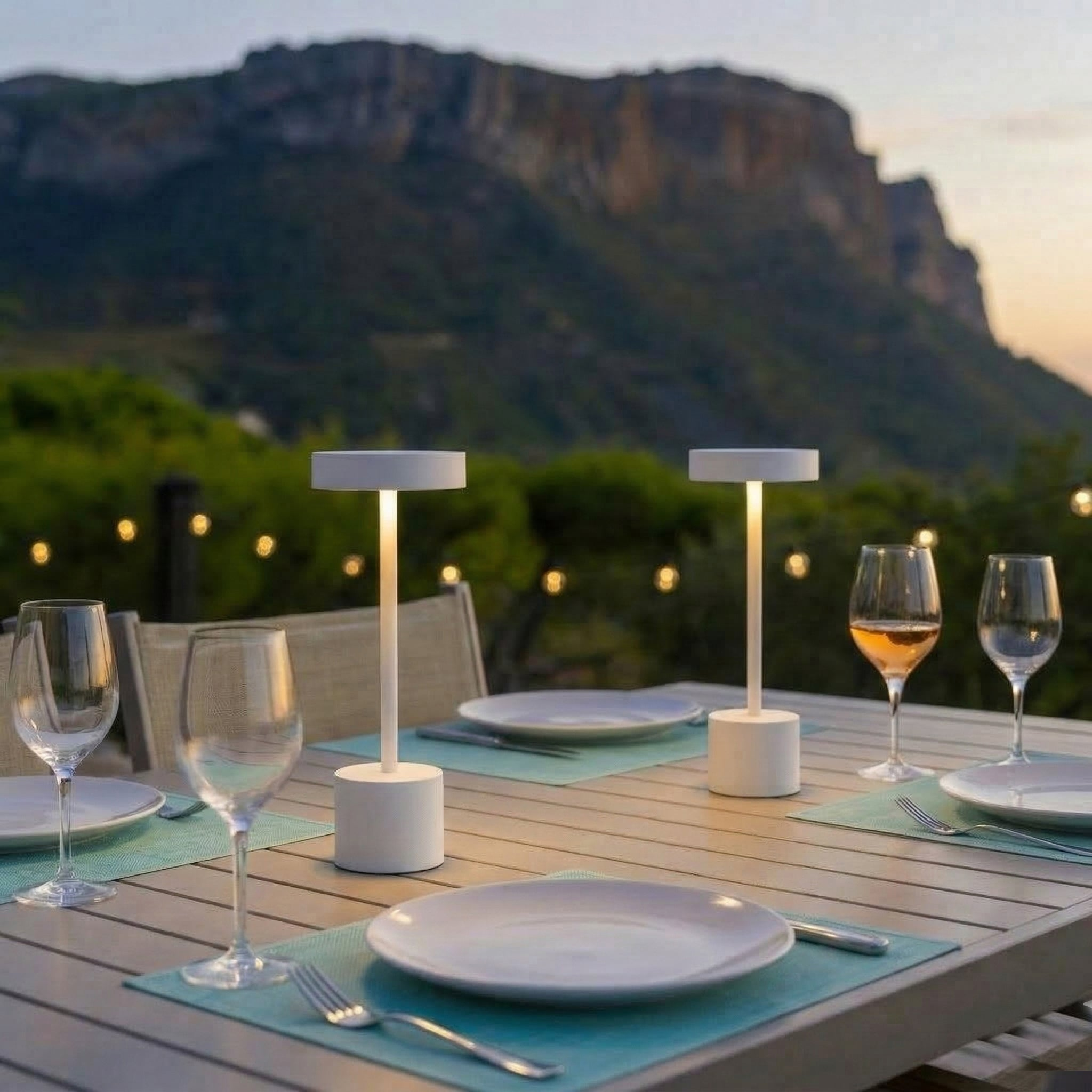 Für ein Abendessen im Freien gedeckter Tisch mit Bergen und Lichterketten im Hintergrund.