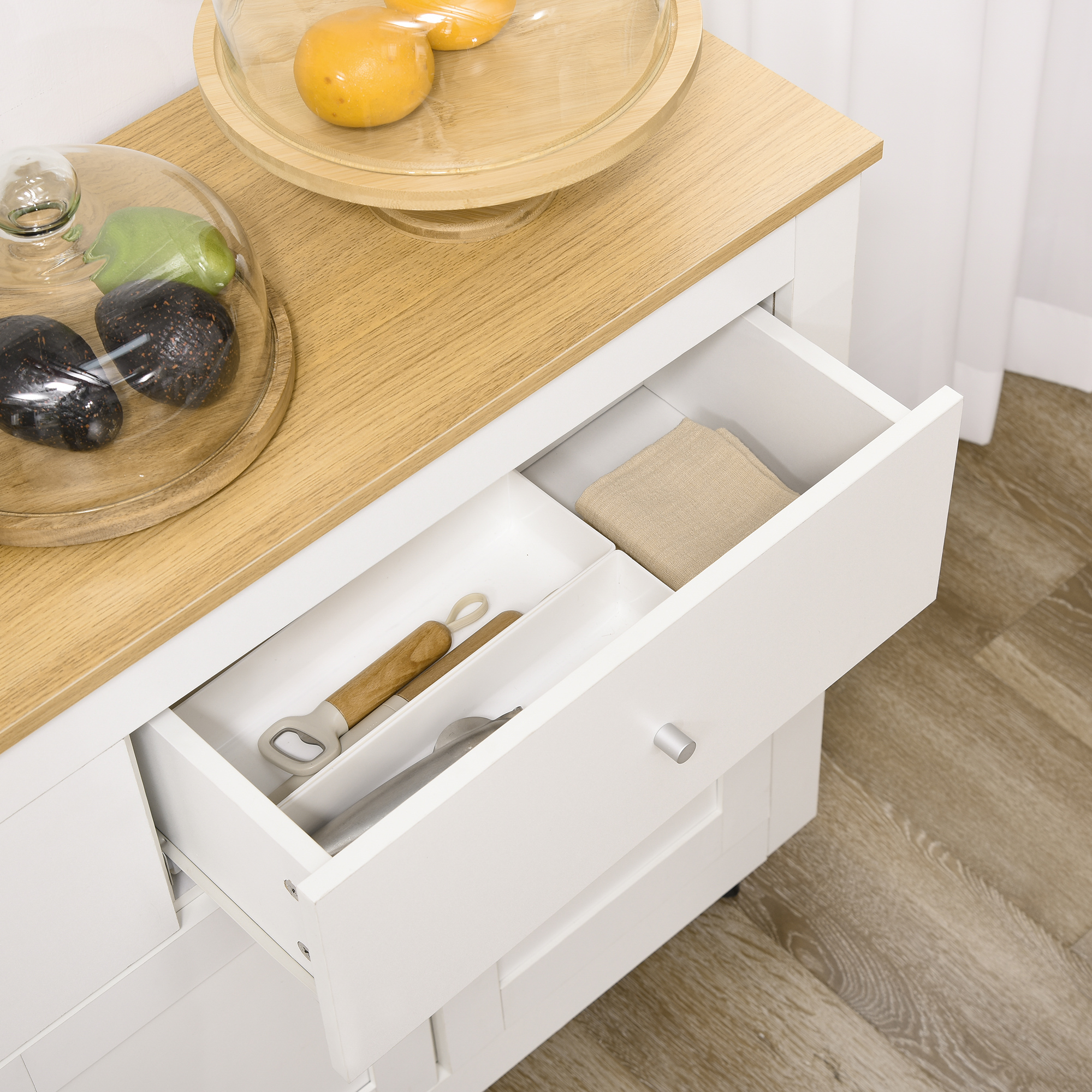 Cajón de cocina abierto con utensilios y fruta bajo campanas de cristal sobre superficie de madera.