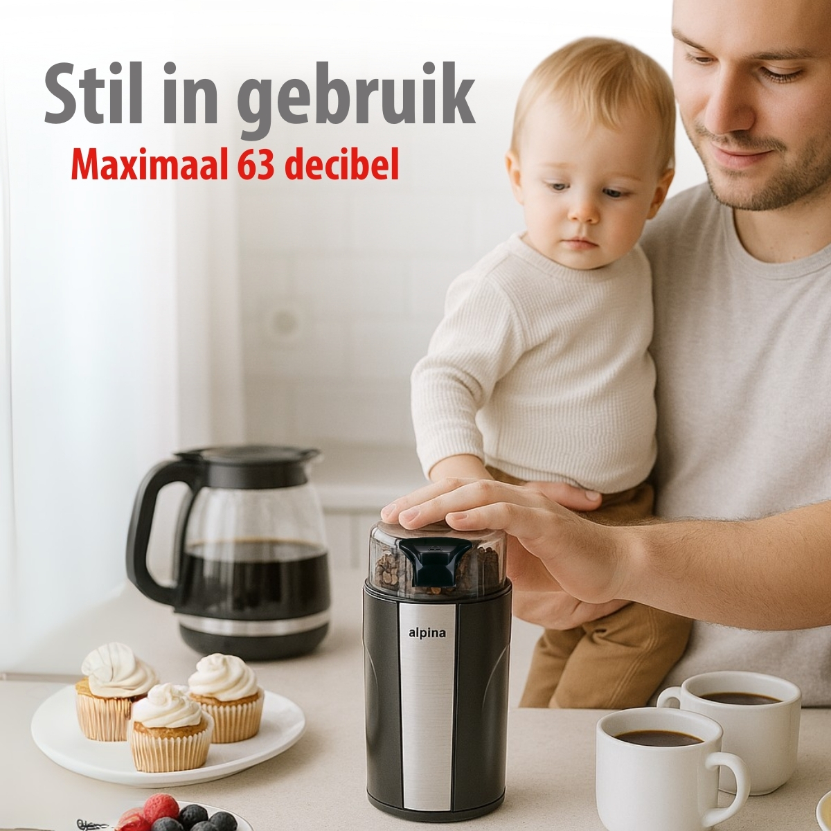 Man holds baby, coffee grinder, cups and cupcakes on a kitchen counter.