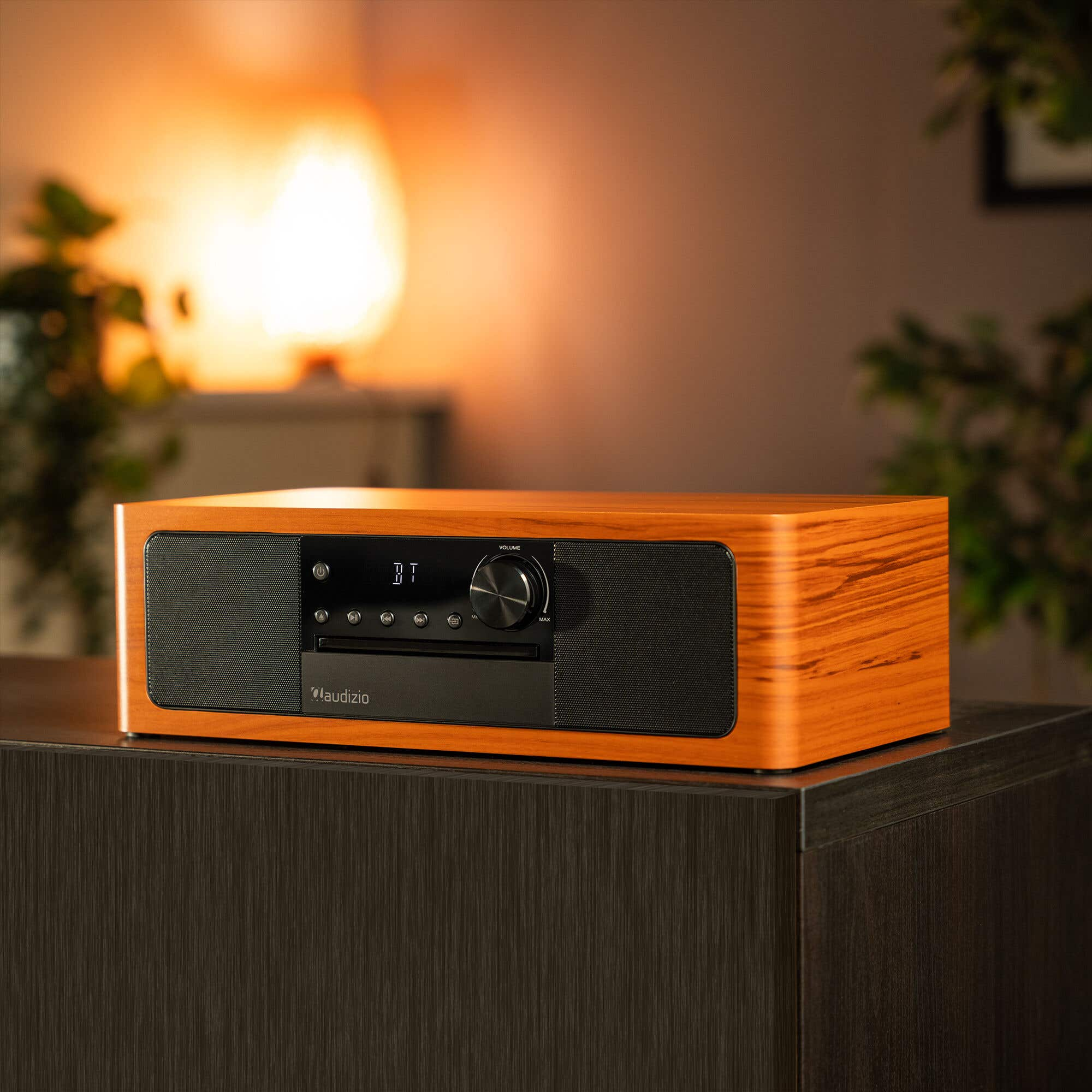 A wooden radio with black speakers and CD slot sits on a dark cabinet. Soft light in background.