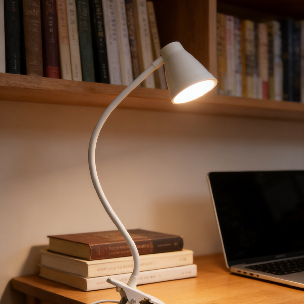 Een witte clip-lamp schijnt op een bureau naast boeken en een laptop.