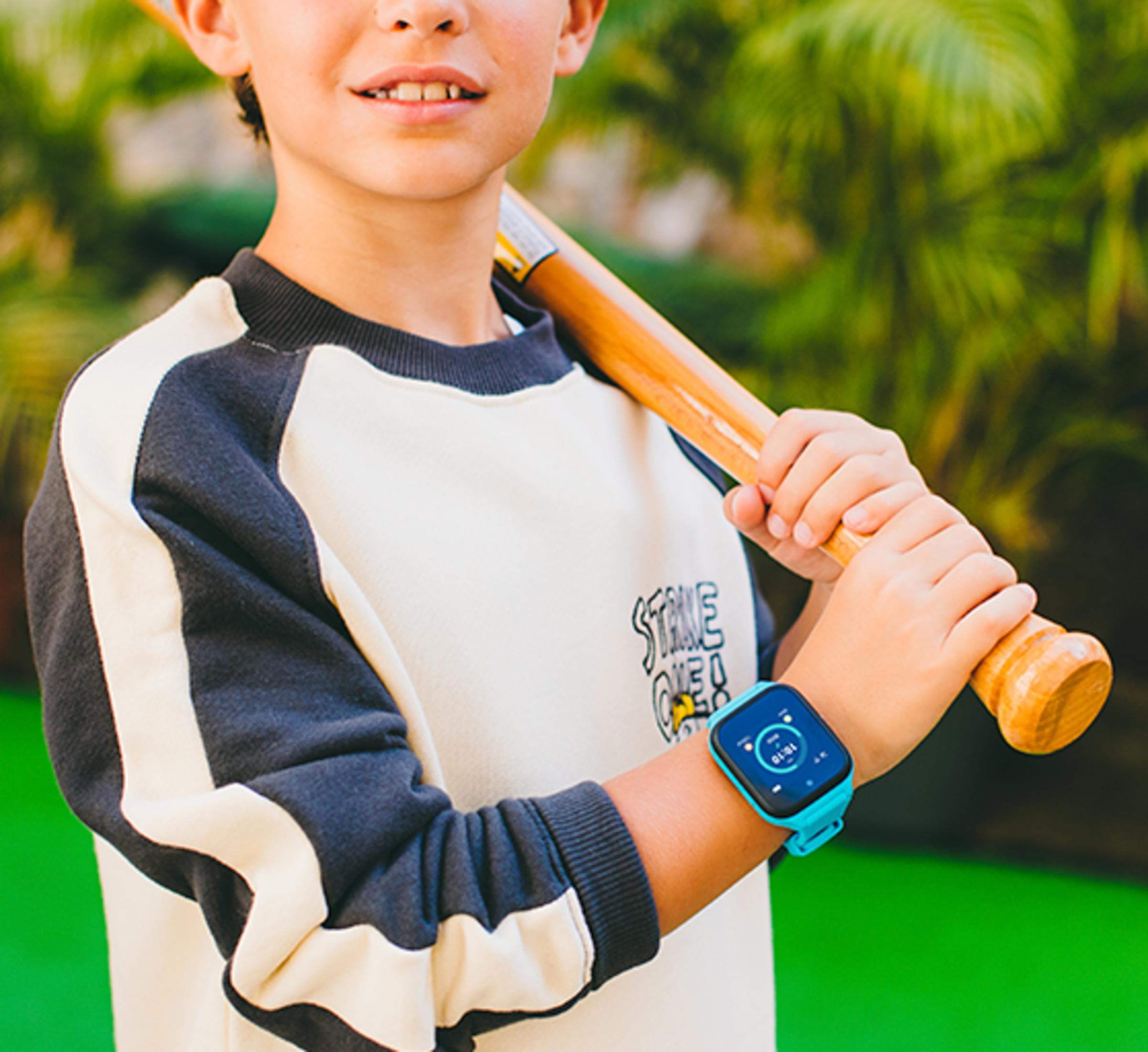 Ein Junge mit Baseballschläger und blauer Smartwatch, im Freien mit grünem Hintergrund.