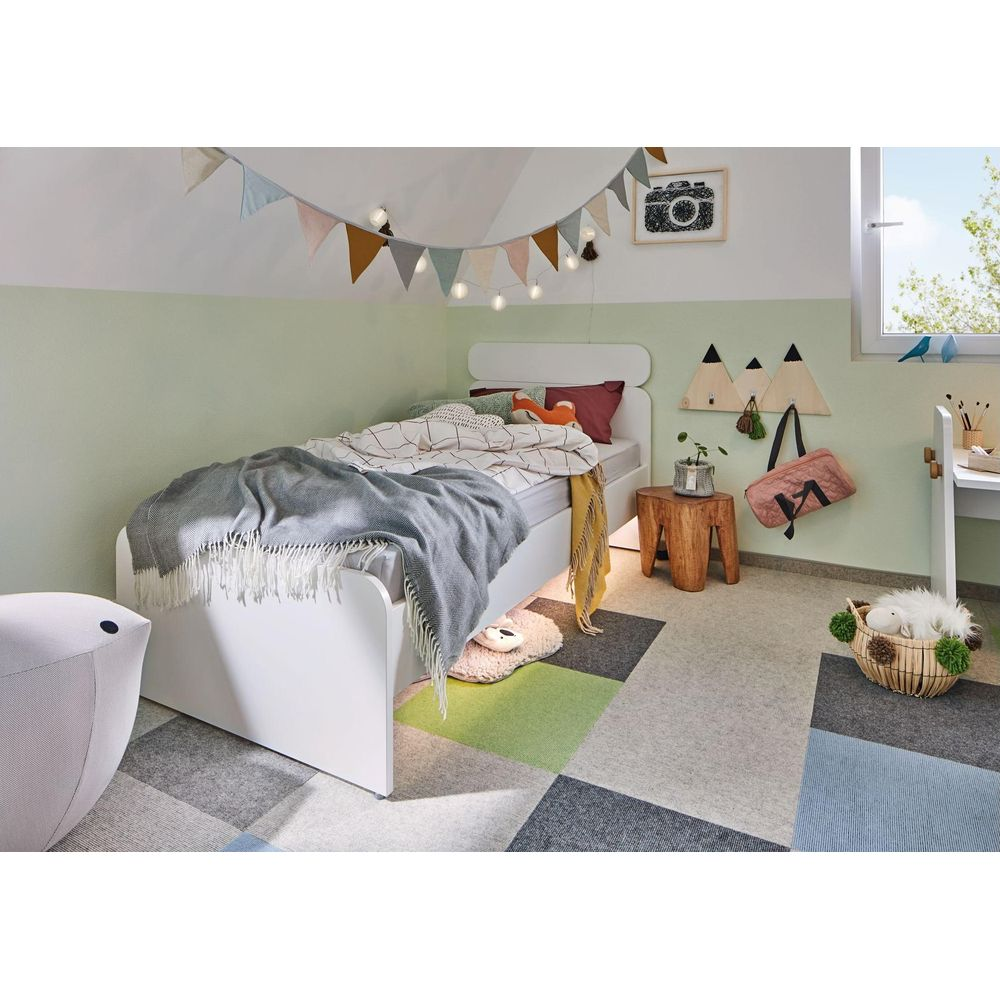 Kinderzimmer mit weißem Bett, buntem Teppich, Deko und Fenster.