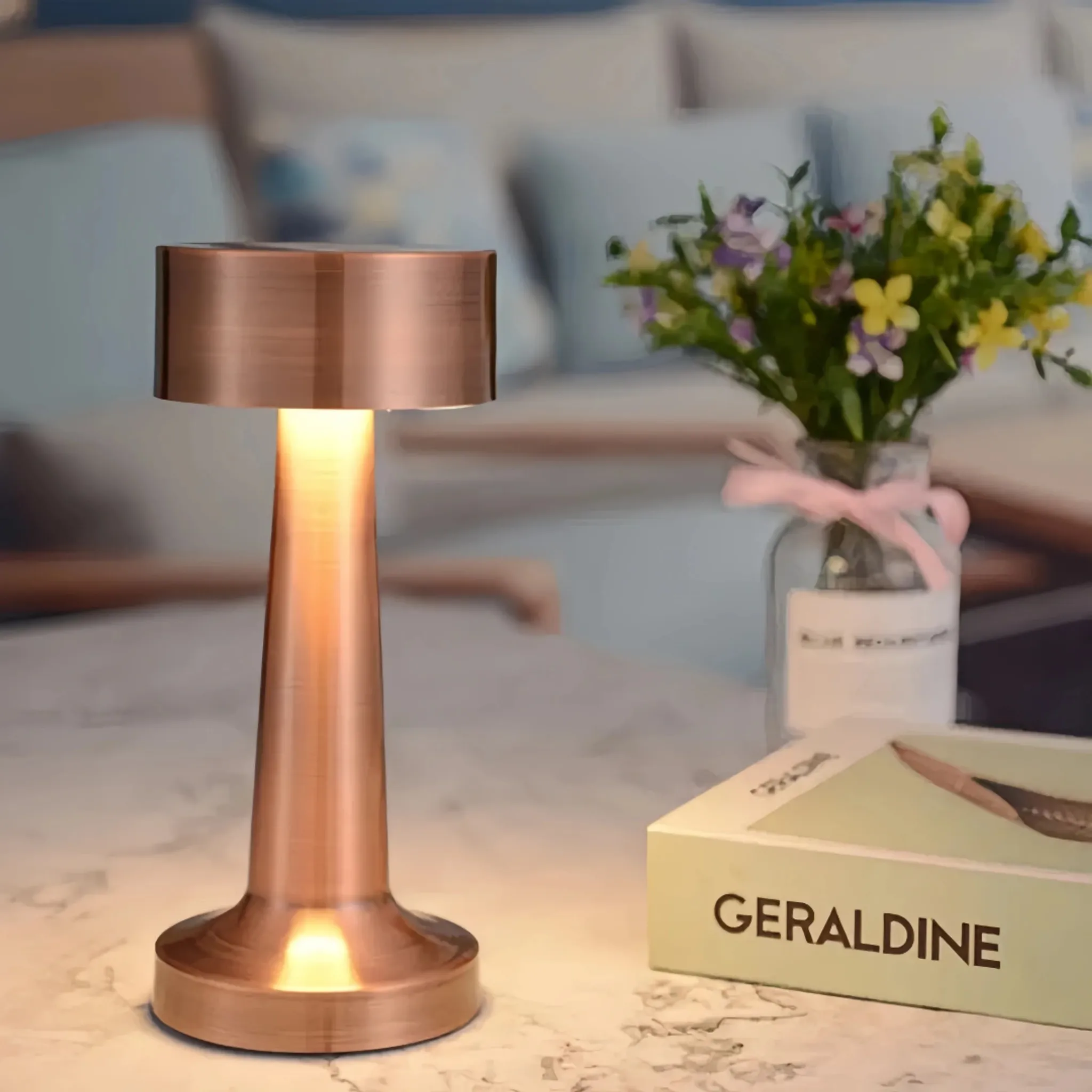 Eine kupferfarbene Lampe leuchtet auf einem Tisch neben Blumen und einem Buch.