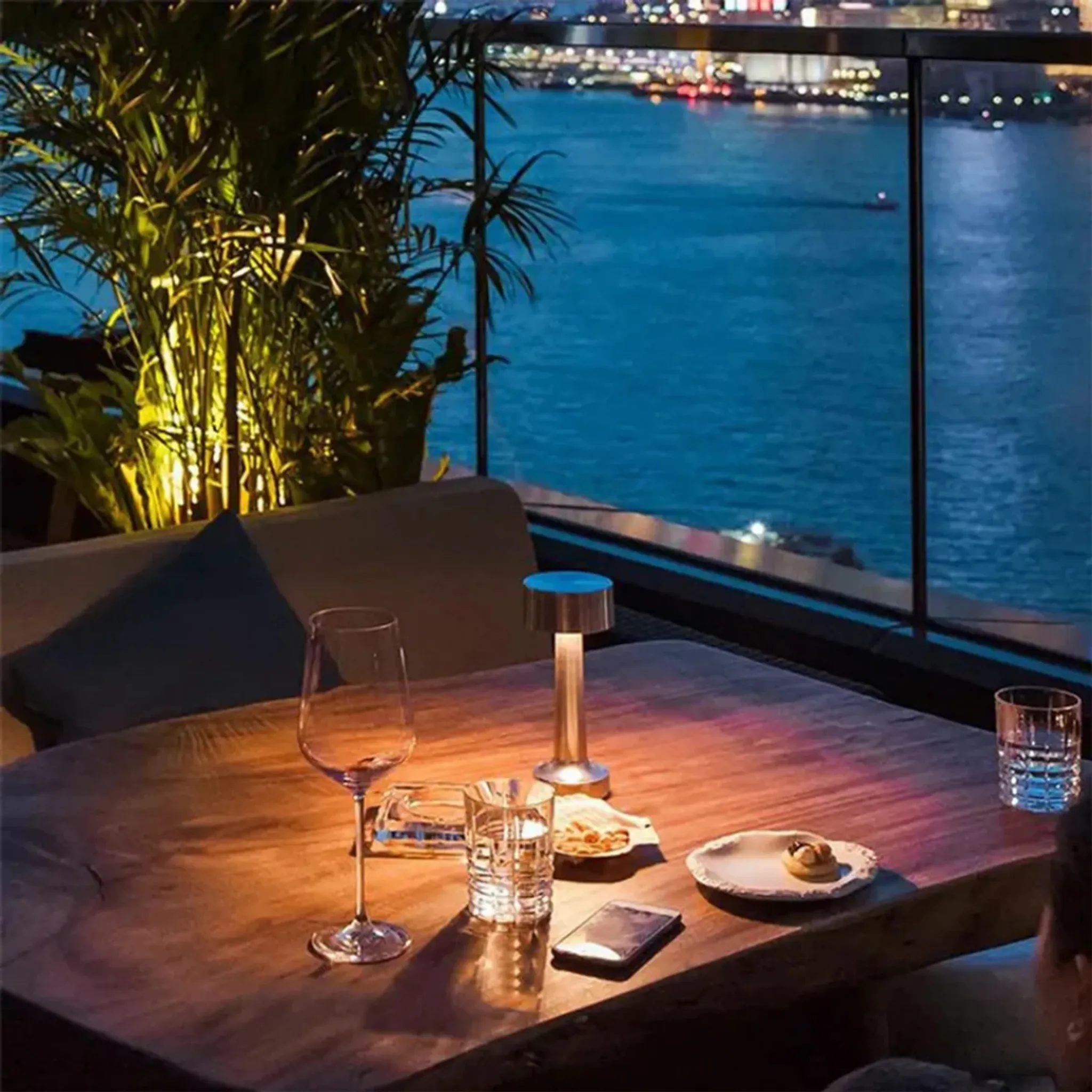 Ein Tisch mit Gläsern, Lampe und Snack auf einer Terrasse mit Meerblick bei Nacht.