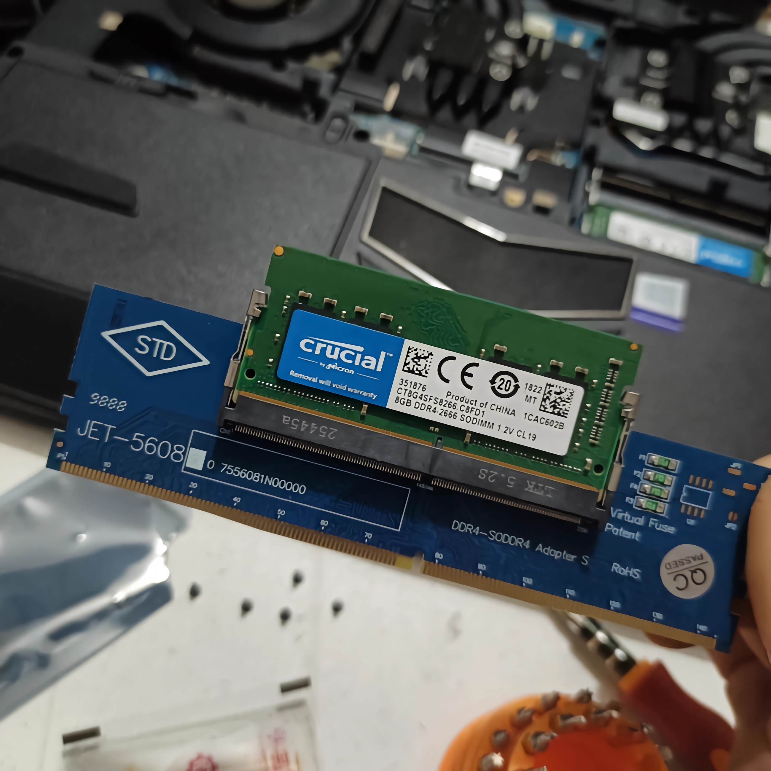 Ein Crucial 8GB DDR4 RAM-Modul auf einem blauen Adapter vor einem geöffneten Laptop.