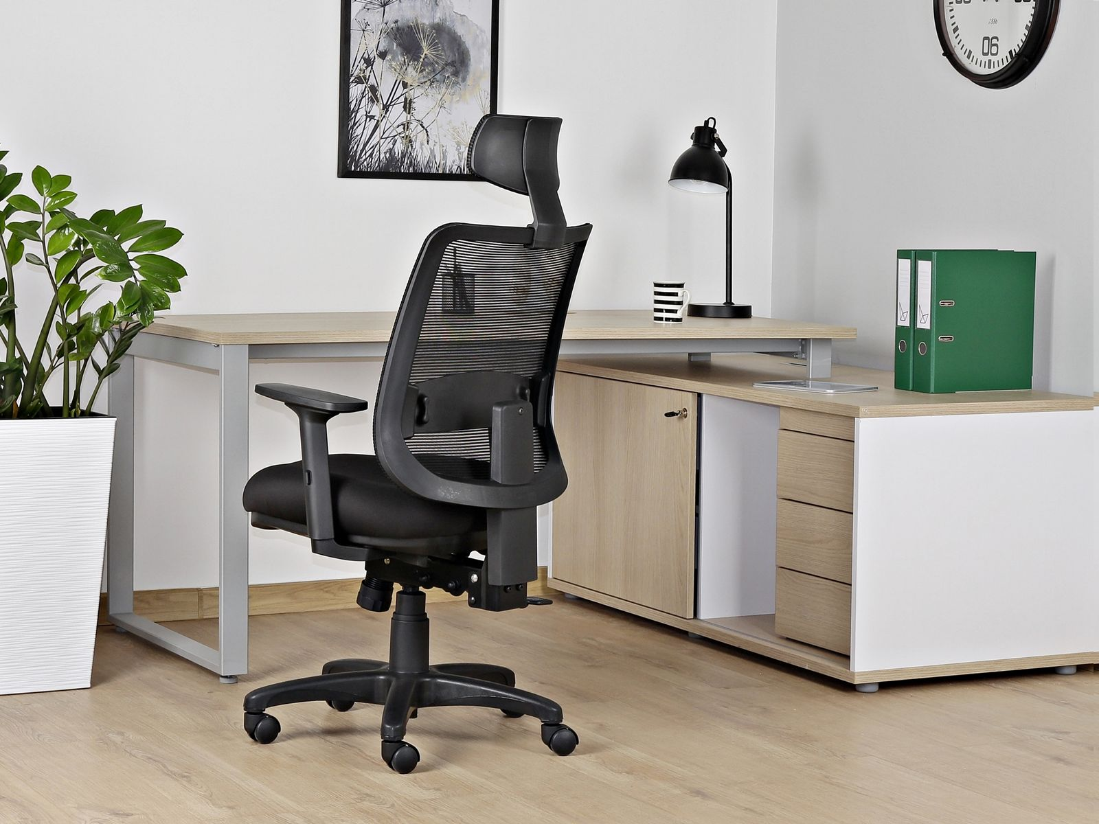 Czarny fotel biurowy stoi przed biurkiem z roślinami, lampą i segregatorami.