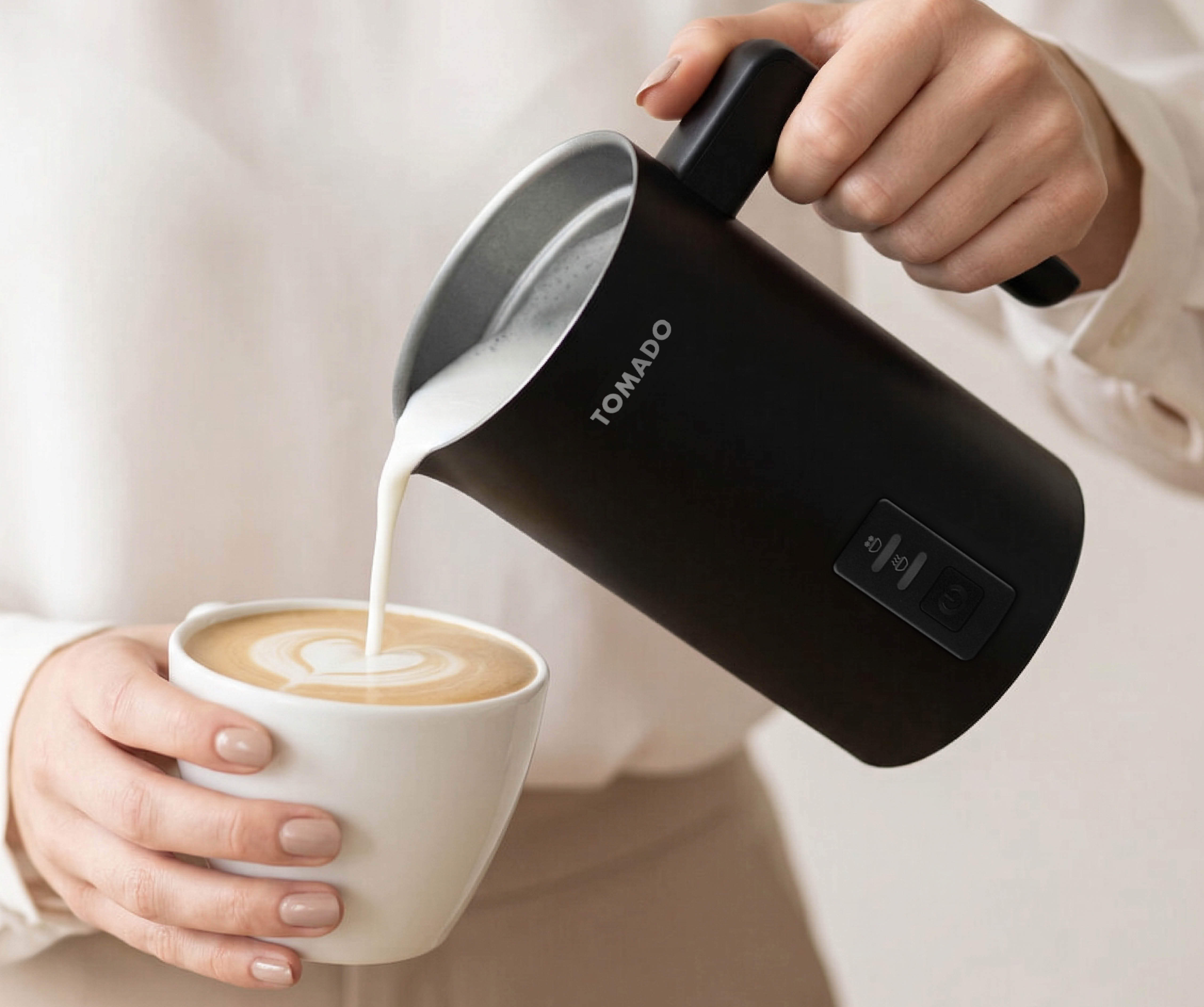 A black milk frother jug pours foam into a white coffee cup with latte art.