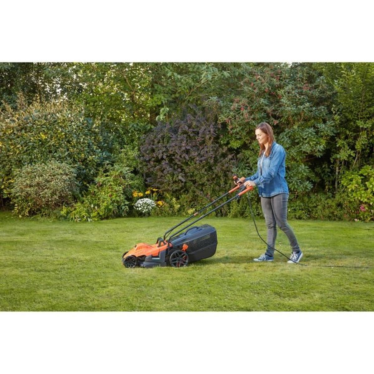Eine Frau in Jeans mäht mit einem orangefarbenen Rasenmäher den Rasen.