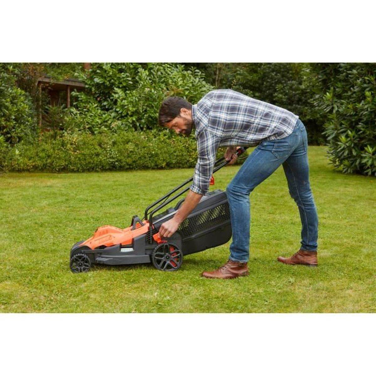 Mann in kariertem Hemd und Jeans stellt einen orange-schwarzen Rasenmäher auf grünem Rasen ein.
