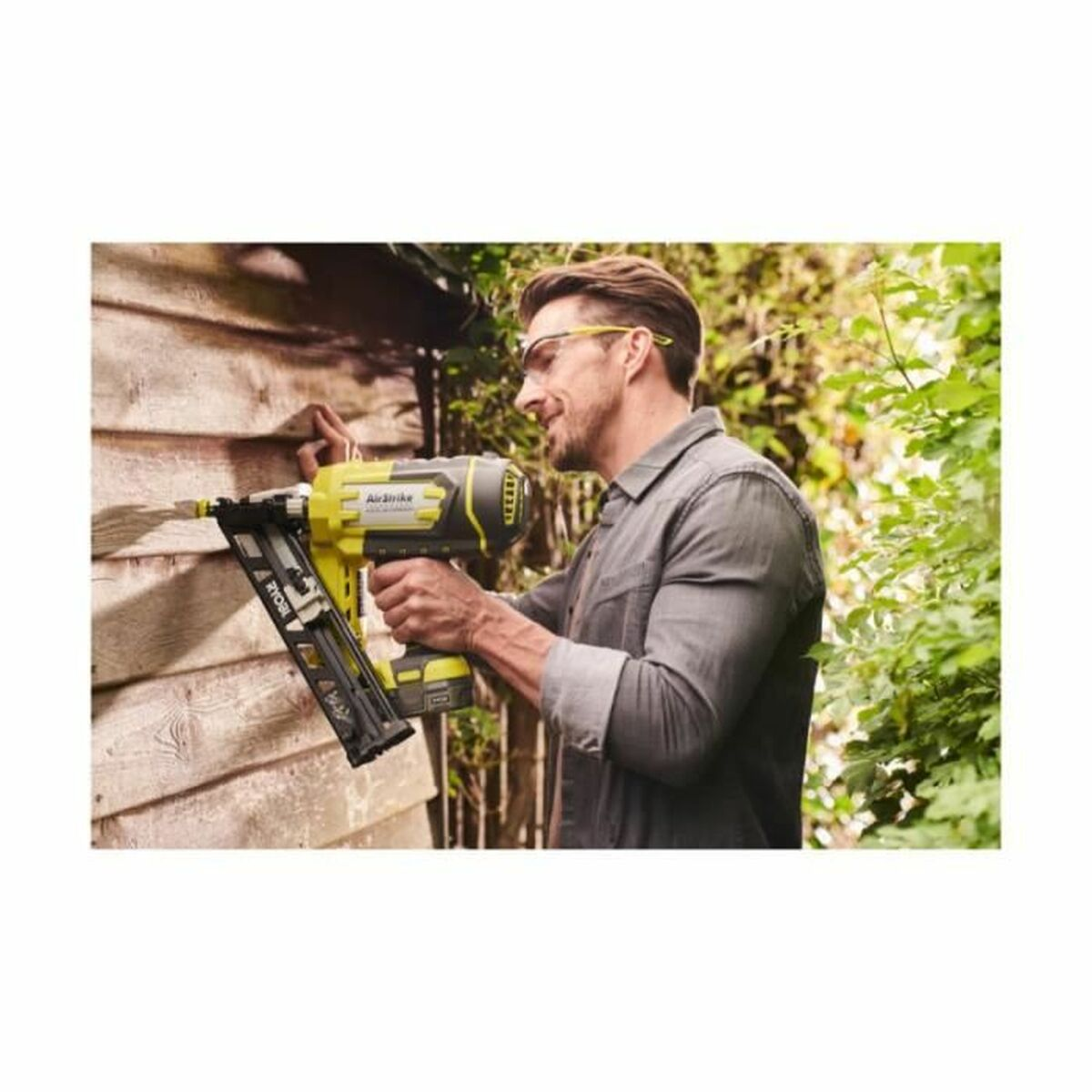 Mann mit Nagelpistole befestigt Holz an einer Wand, Garten im Hintergrund.