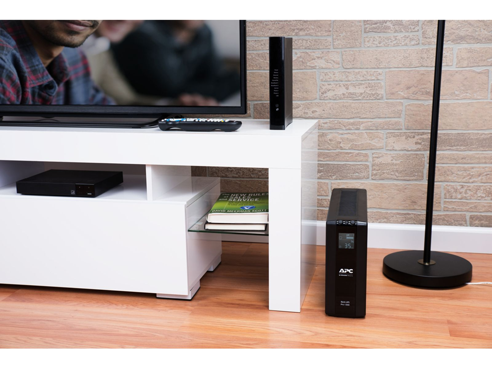 Ein Mann schaut fern. Ein weißes TV-Möbel mit Geräten, Büchern und einer Lampe.