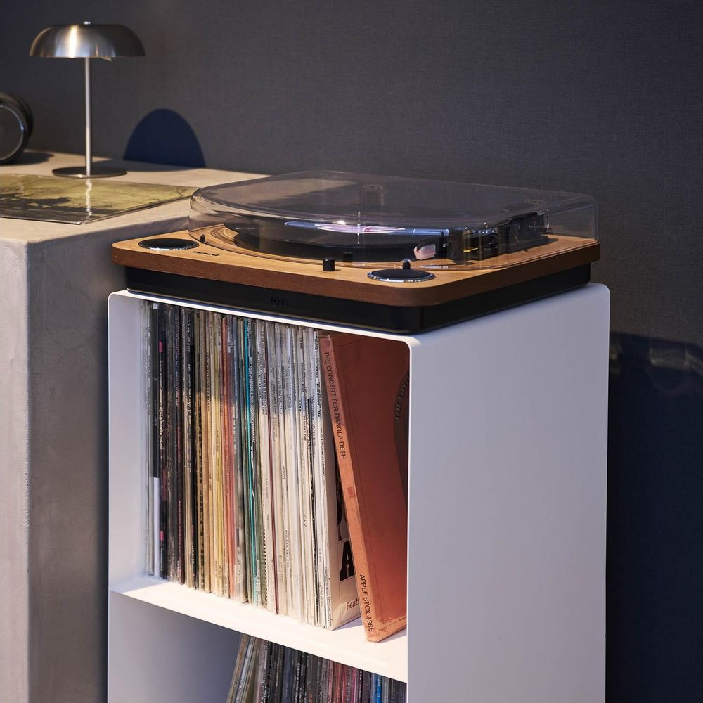 Ein Plattenspieler steht auf einem weißen Regal voller Schallplatten. Links steht eine Lampe.