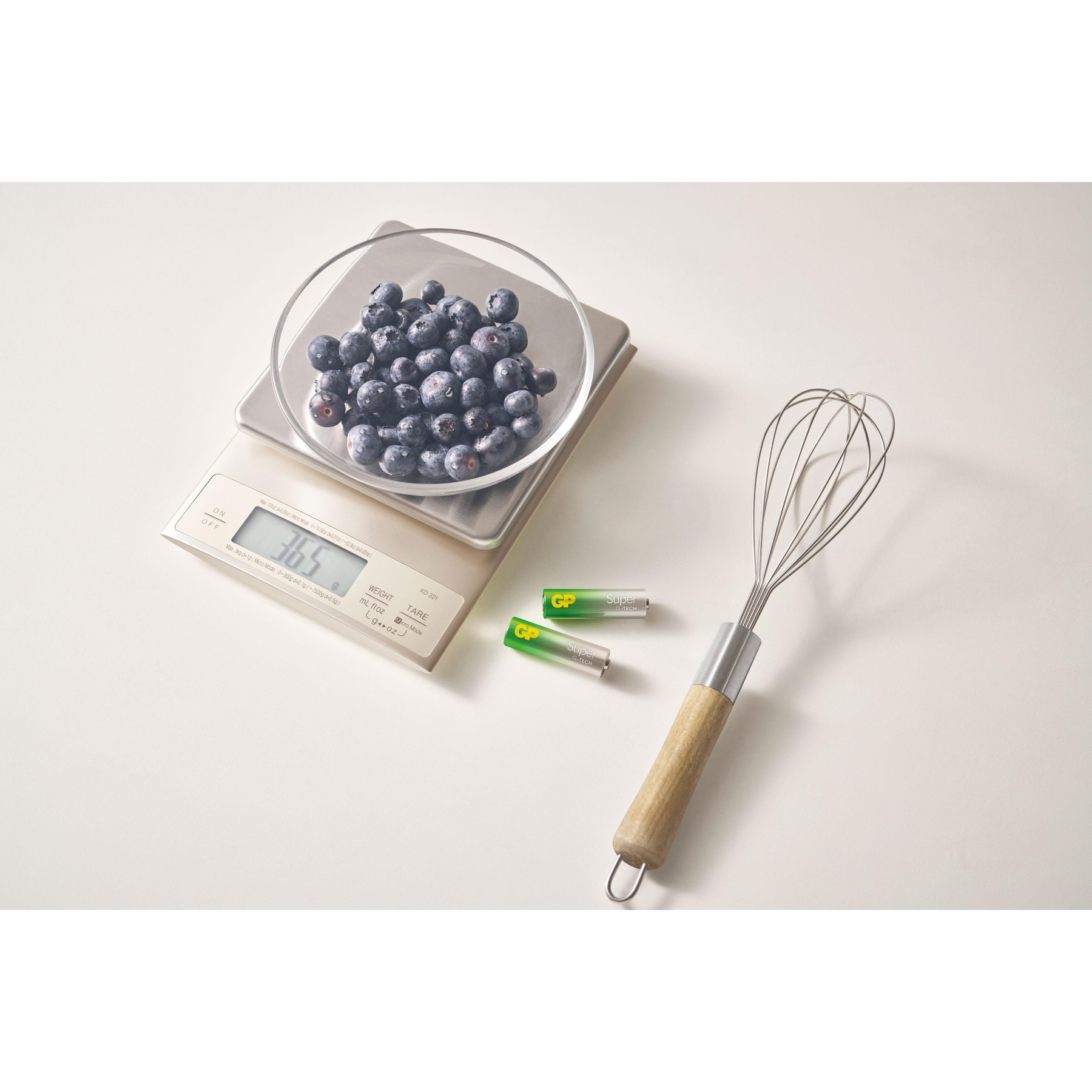 Waage mit Blaubeeren, zwei Batterien und Schneebesen auf weißem Hintergrund.
