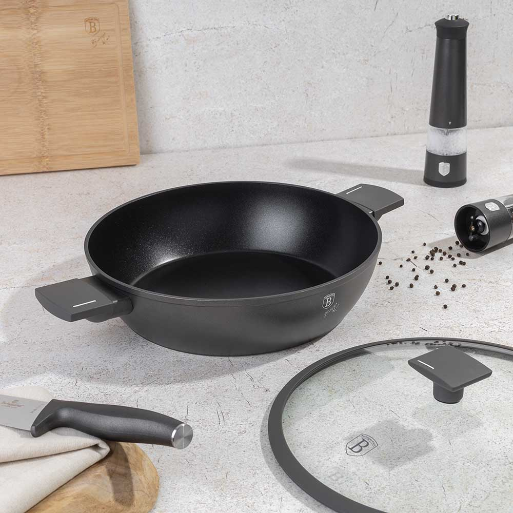 Eine schwarze Bratpfanne mit Deckel, Messer, Holzbrett und Gewürzmühlen auf Marmor.