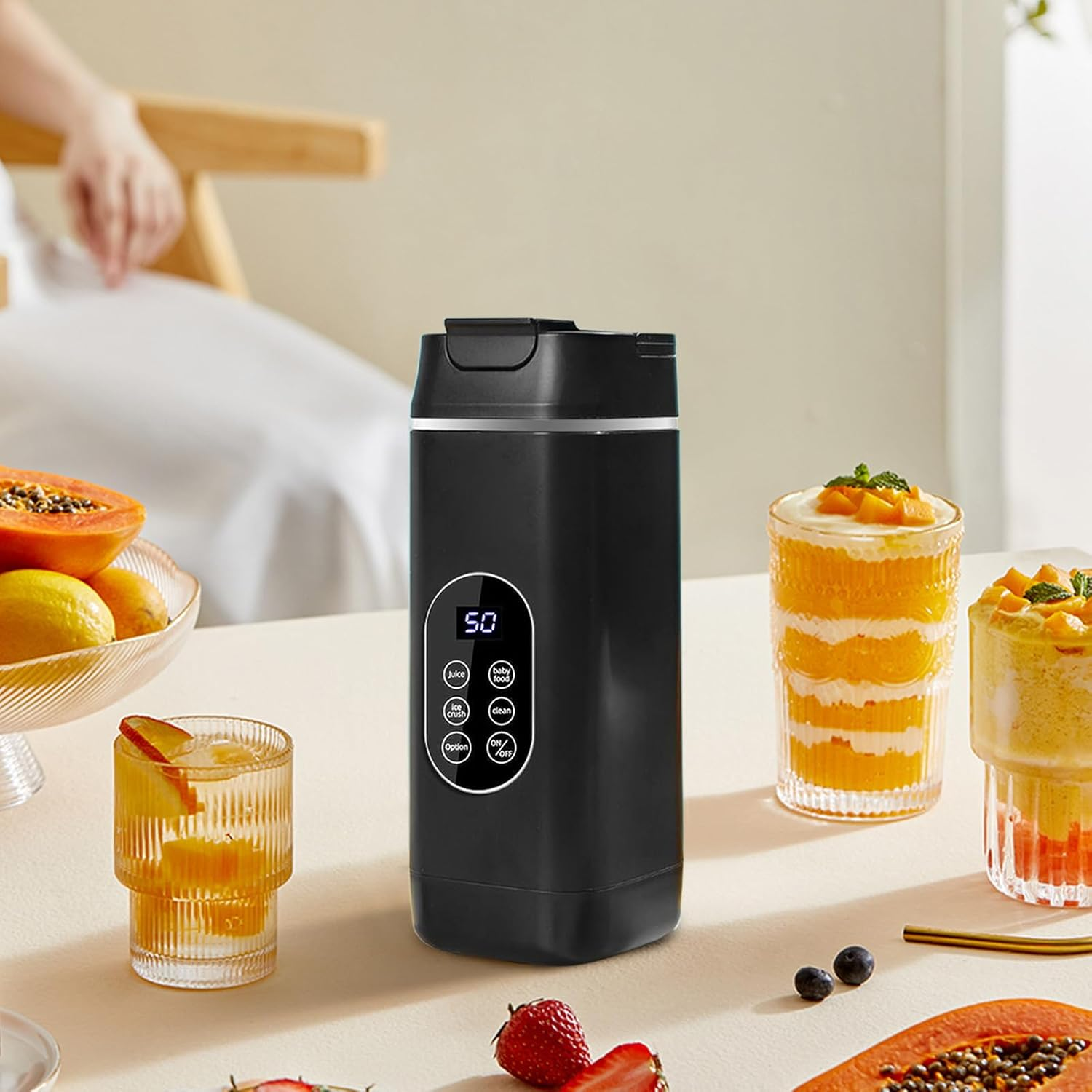 Schwarzer Mixer auf einem Tisch mit Obst, Getränken und einer Person im Hintergrund.