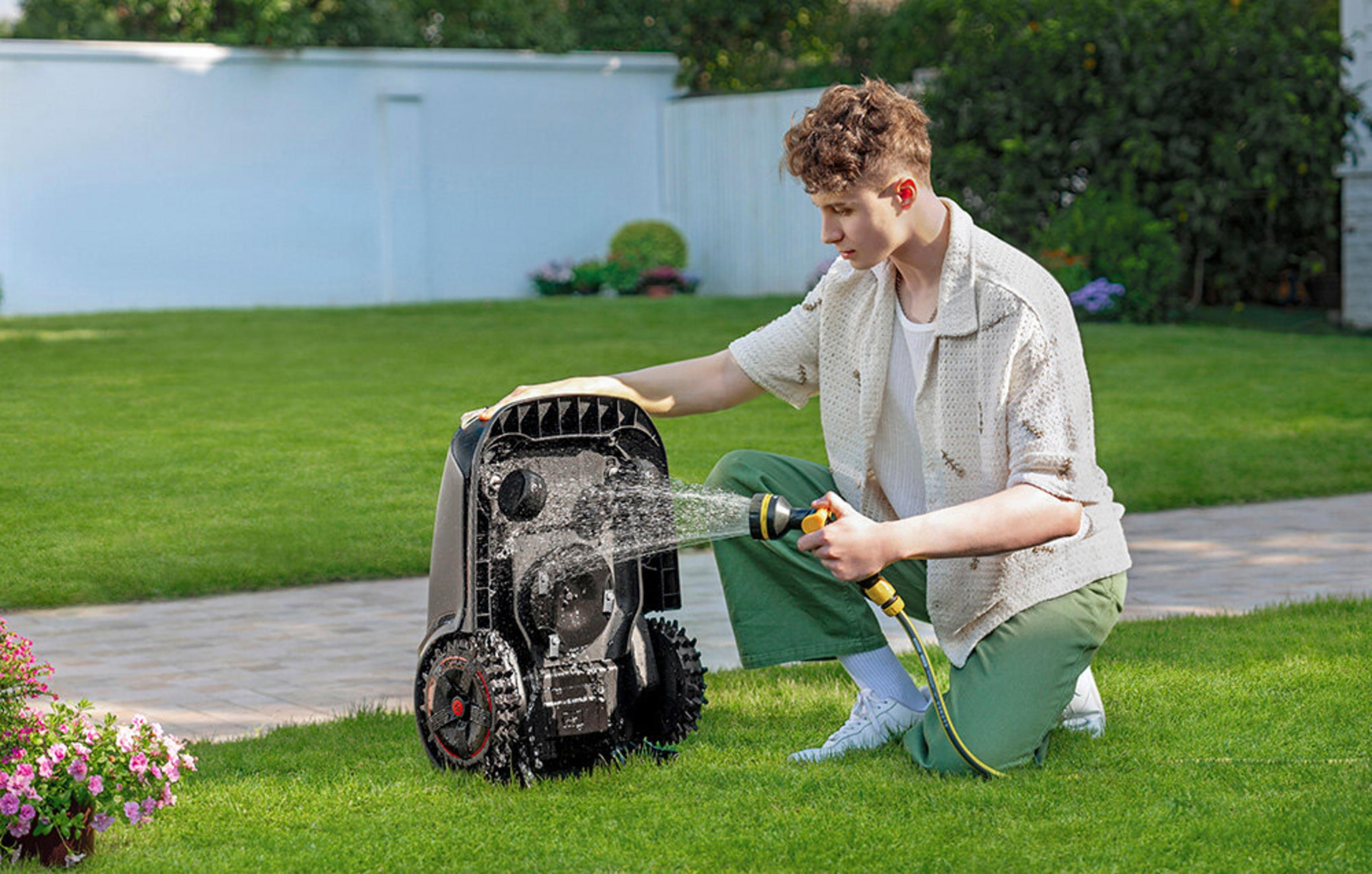 Mężczyzna spryskuje robot koszący trawnik wężem na trawniku.