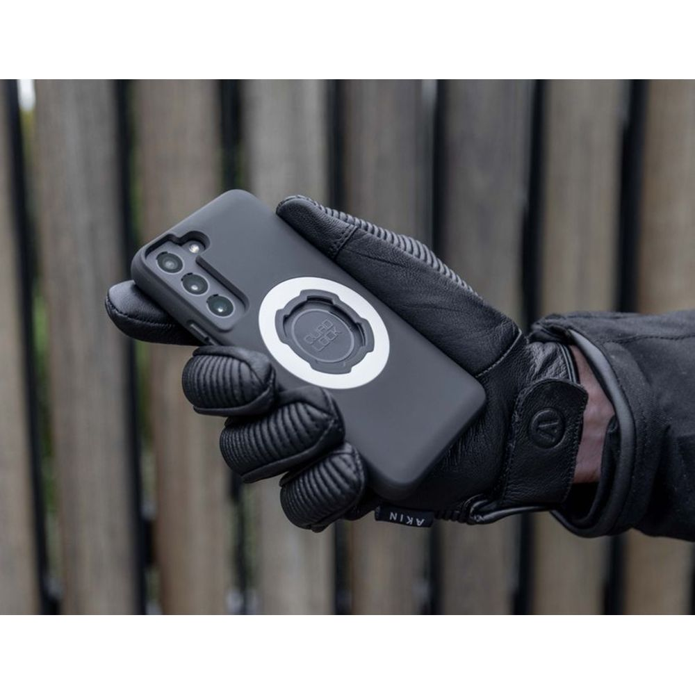 Eine Hand mit schwarzem Handschuh hält ein Smartphone mit Quad Lock Halterung vor einem Holzzaun.