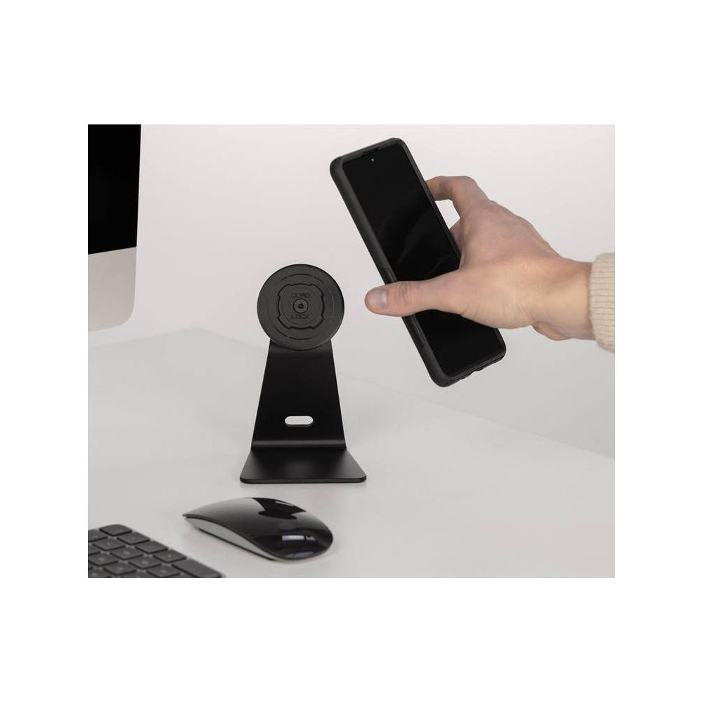Eine Hand hält ein Handy in der Nähe eines schwarzen Handyständers auf einem weißen Schreibtisch mit Maus und Tastatur.