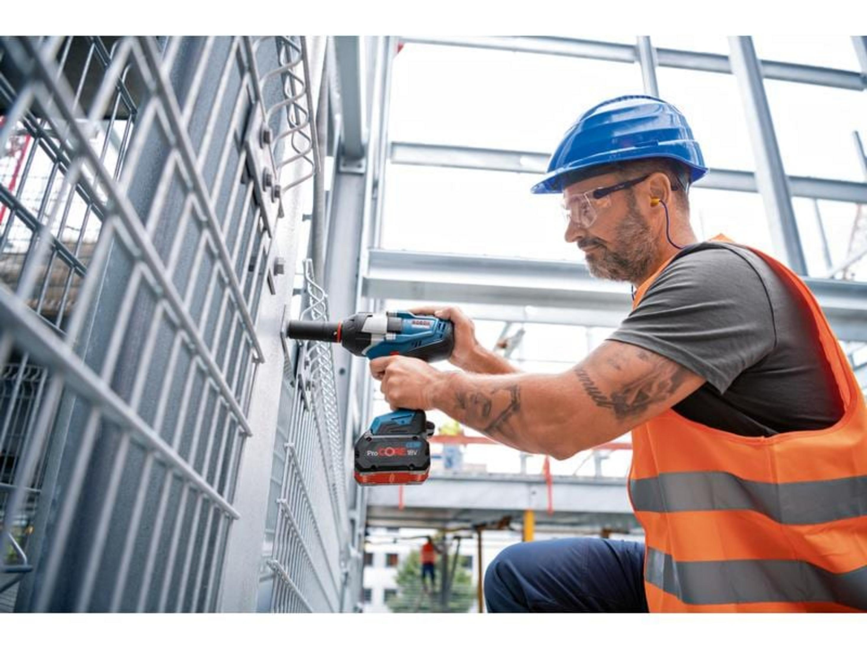 Mann in blauem Helm und orangefarbener Weste benutzt Akkuschrauber an Metallzaun.