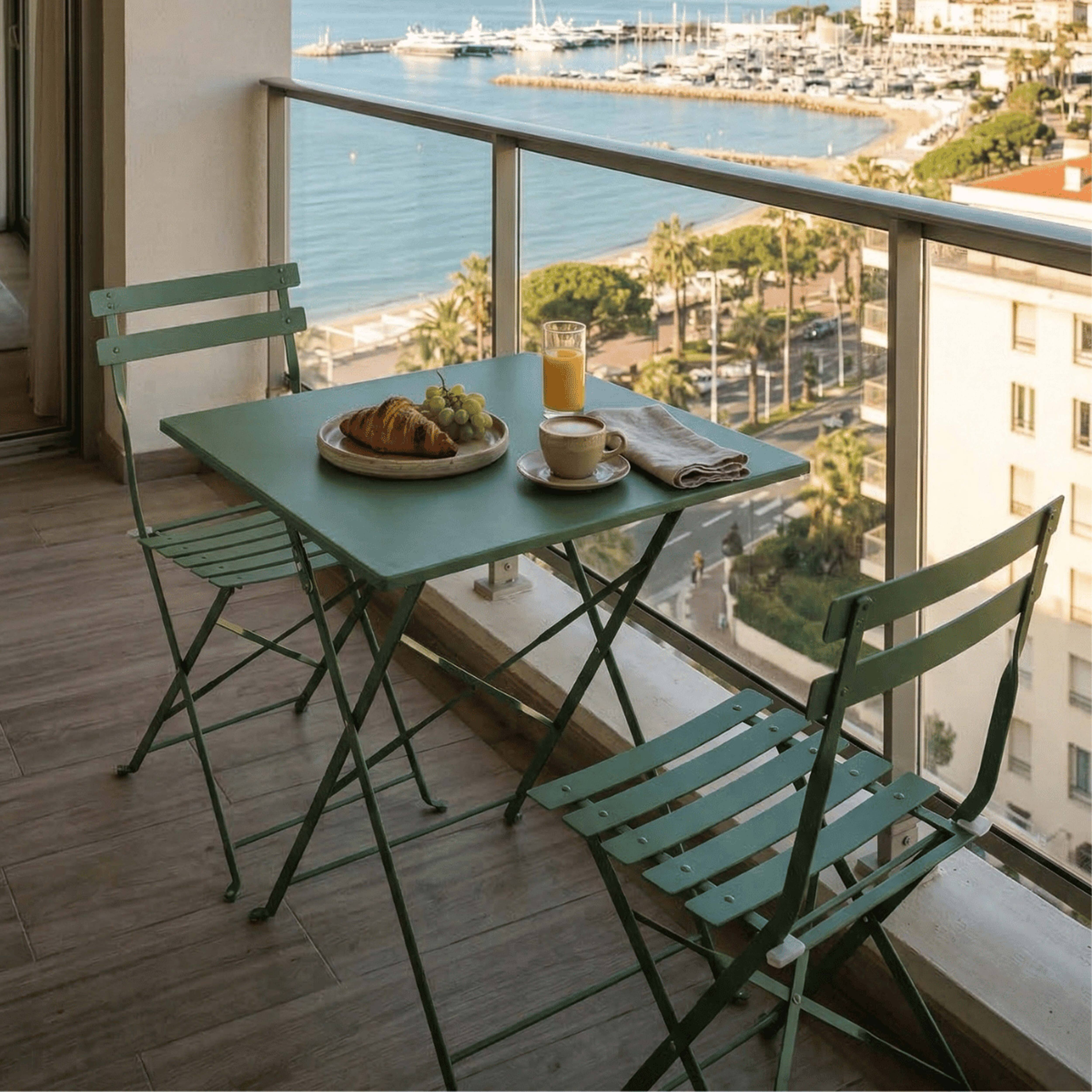 Ein Balkon mit einem grünen Tisch und zwei Stühlen, gedeckt mit Frühstück, mit Blick auf eine Bucht mit Booten und Gebäuden.