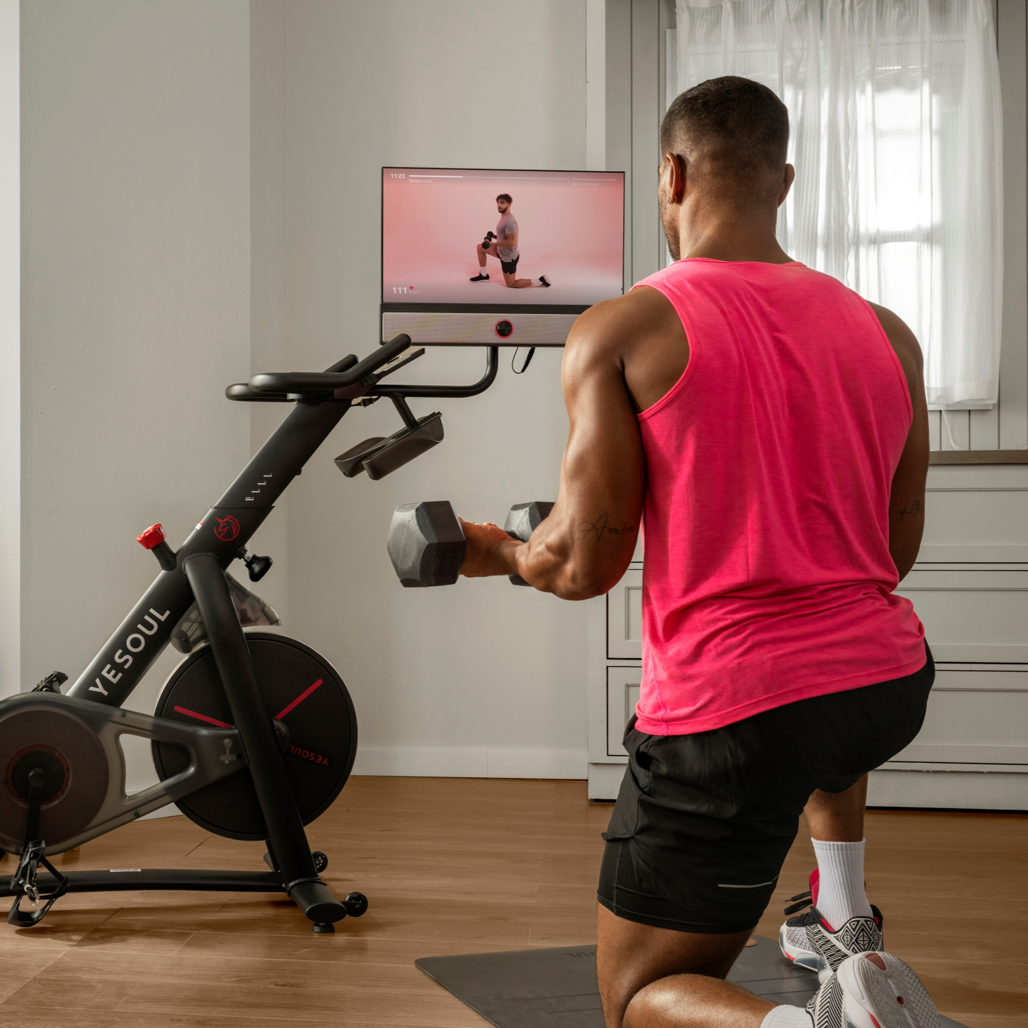 Ein Mann in einem pinken Tanktop trainiert mit Hanteln vor einem Fitnessrad und einem Bildschirm.