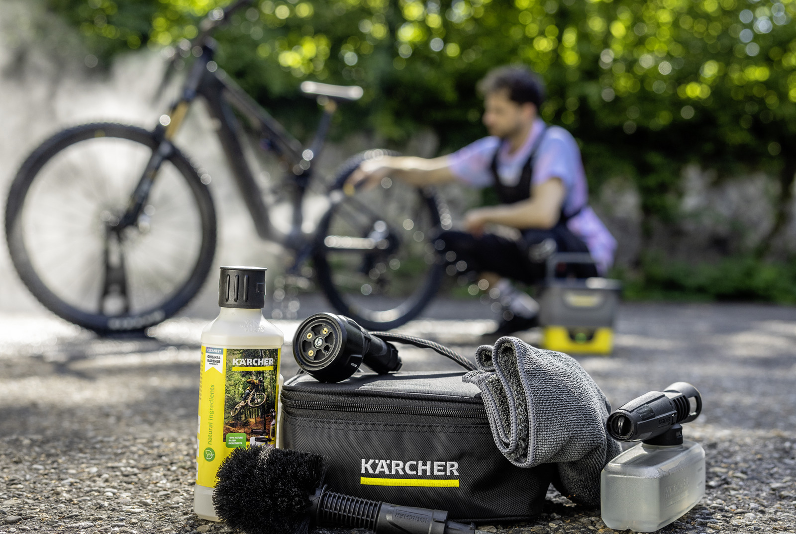 Fahrradreinigungsmittel, Bürste, Tuch, Sprühkopf und Kärcher-Tasche vor einem Fahrrad und Mann.